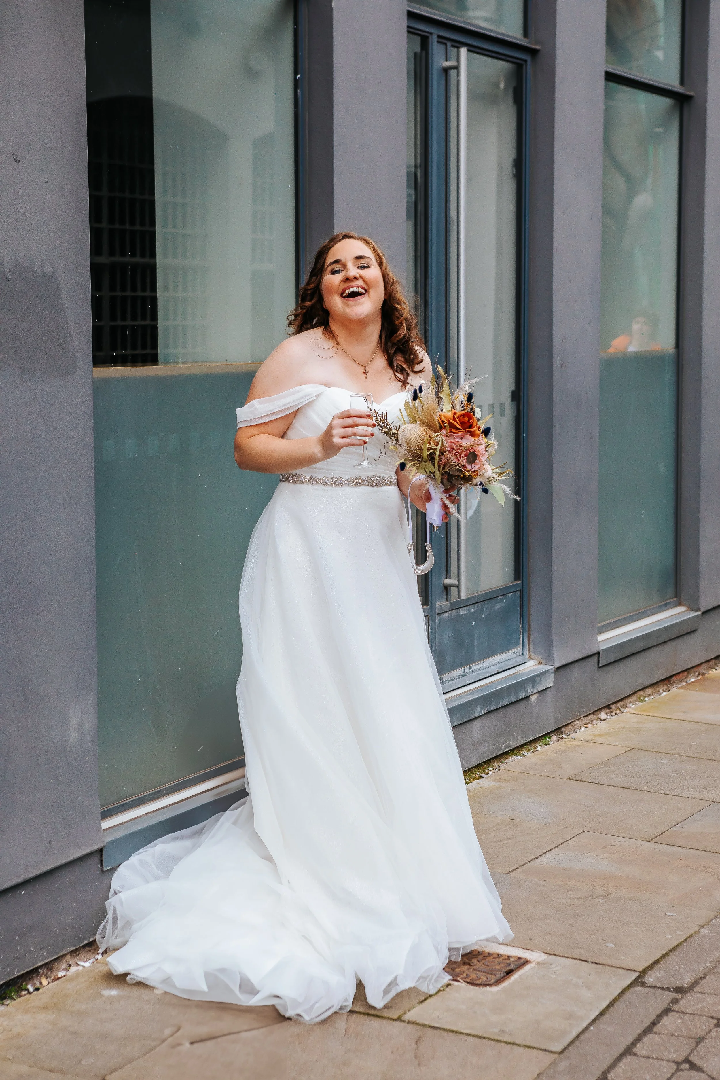 A bride in a white off-shoulder wedding dress holding a bouquet and a glass, smiling and laughing outdoors next to a modern building with large windows.