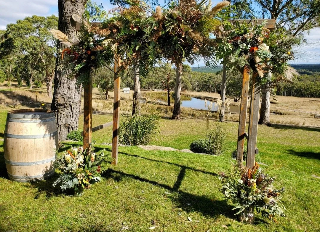 Outdoor wedding arch decorated with flowers and greenery, set in a grassy area with trees and a pond in the background.