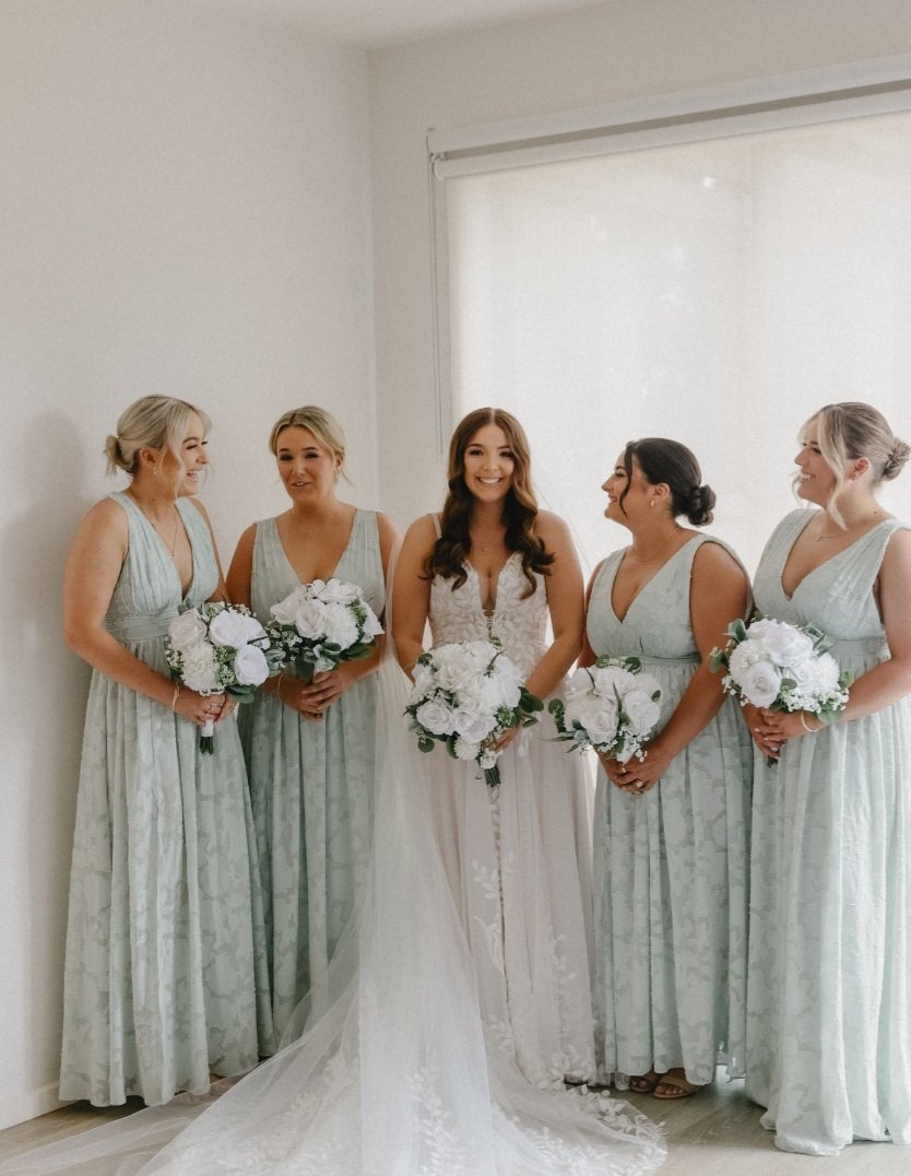 Bride in a white wedding gown and five bridesmaids in light blue dresses holding white bouquets, standing near a window with a white blind.