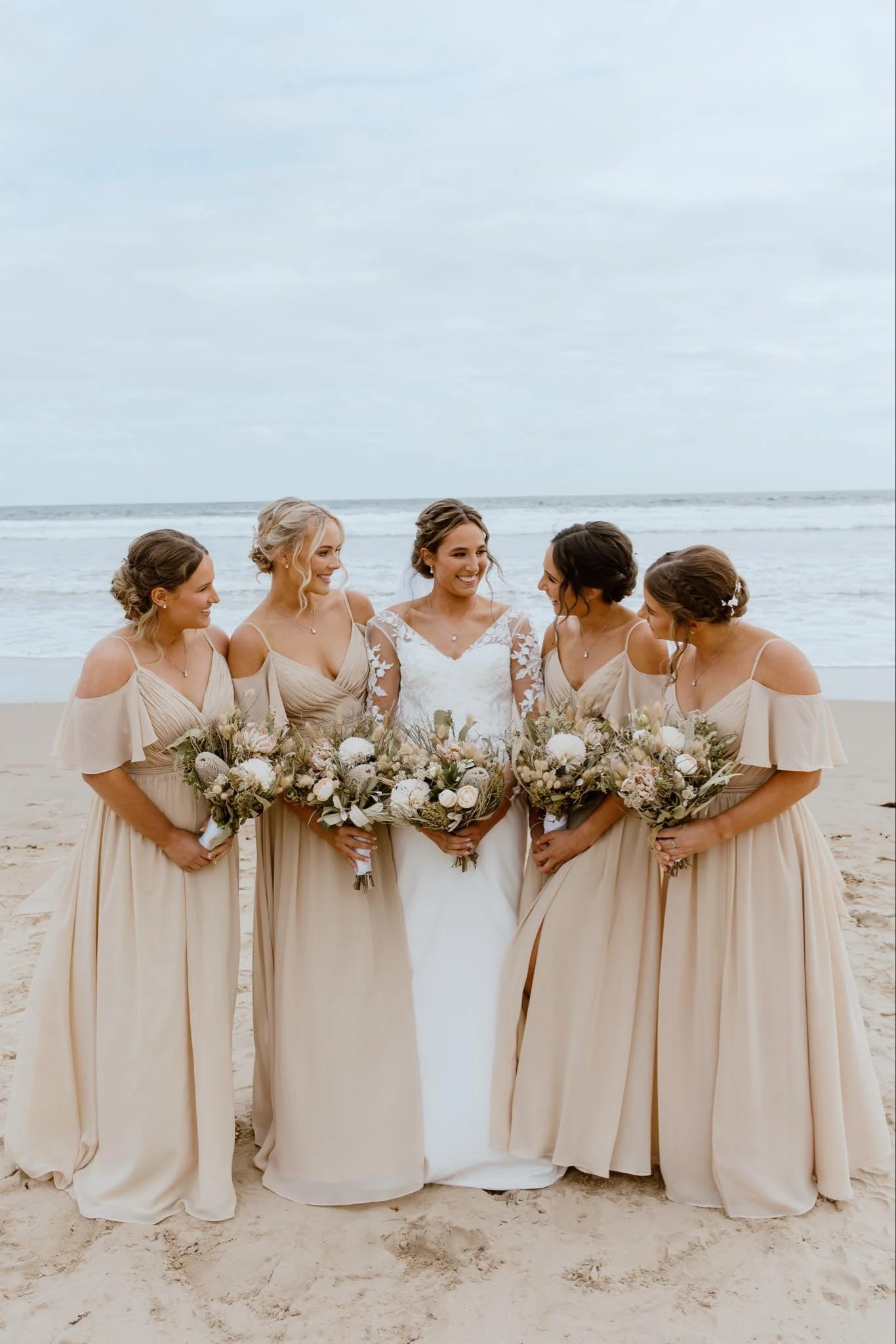 Group of five women on the beach, dressed in formal attire, holding bouquets, smiling and looking at each other.