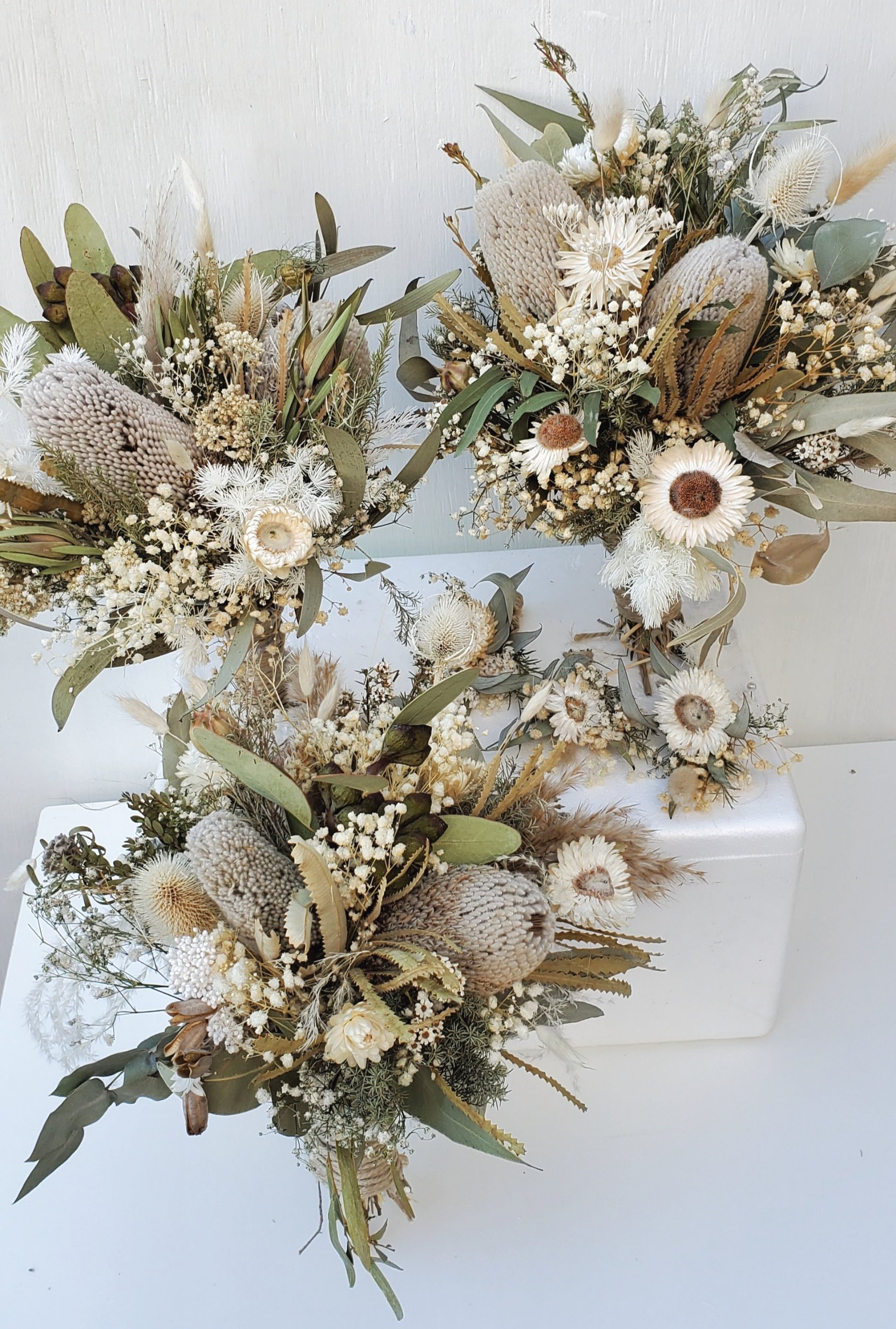 Dried flower arrangements with white and beige flowers, green leaves, and beige or brown accents, placed on a white surface against a light background.