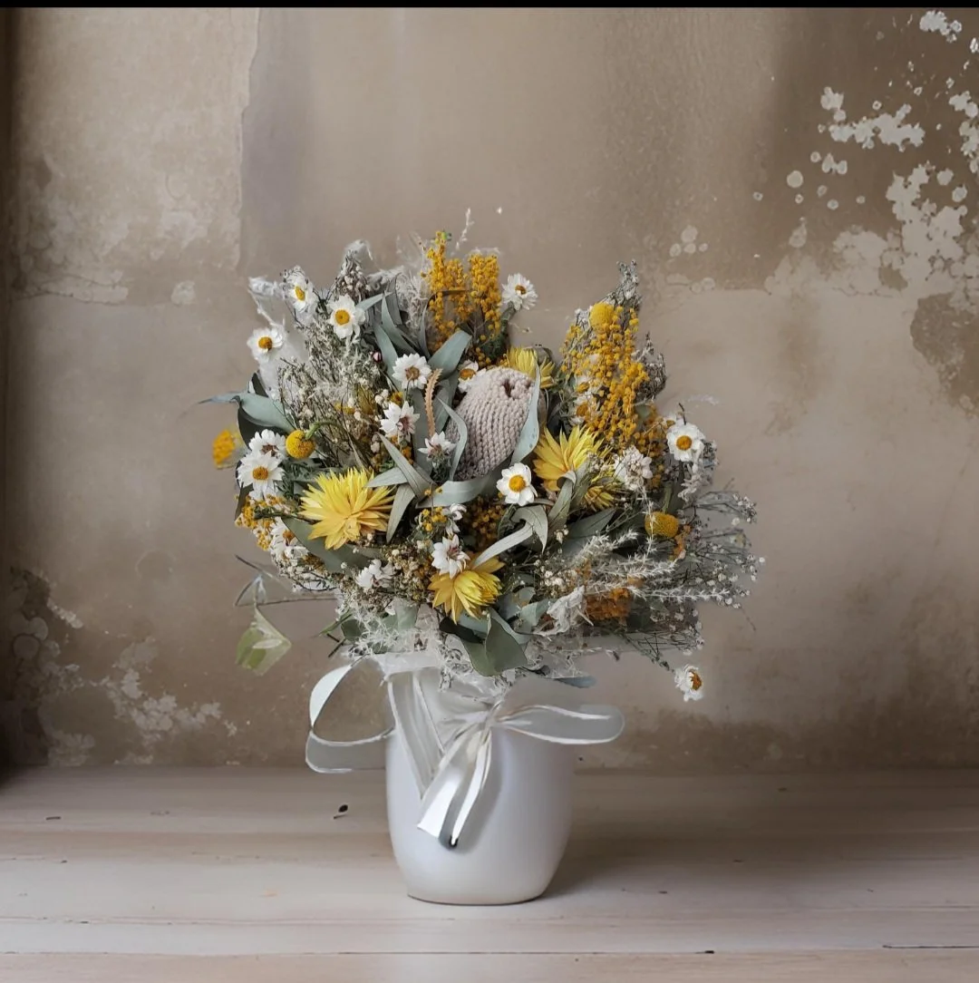 A bouquet of yellow, white, and gray dried flowers in a white vase, with a small stuffed owl among the flowers, and tied with a white ribbon.