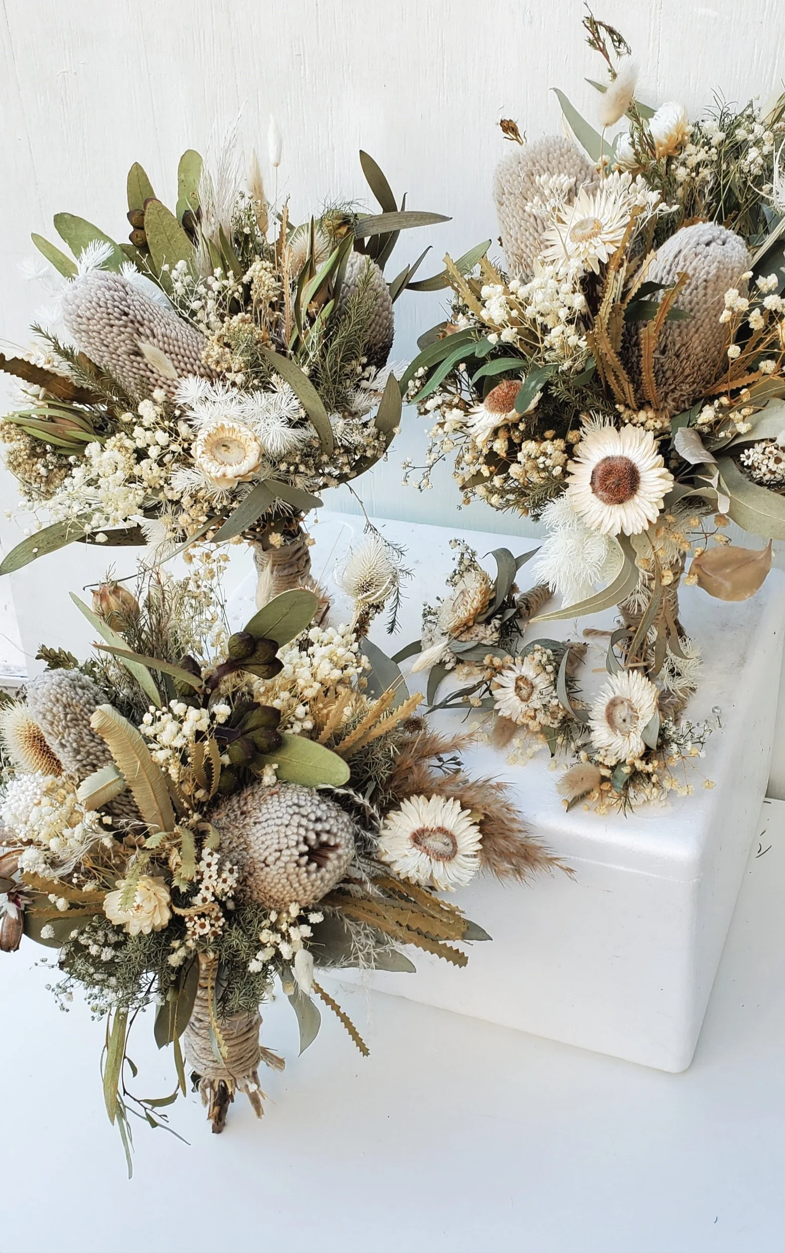 Three dried flower arrangements featuring white, beige, and green foliage, placed on a white surface against a white wall.