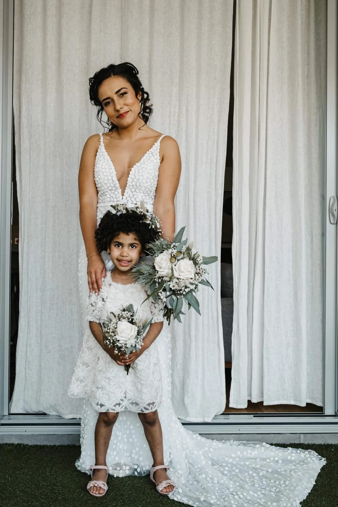 A woman in a white wedding dress, standing behind a young girl in a white lace dress, both holding white floral bouquets, with a curtain background.