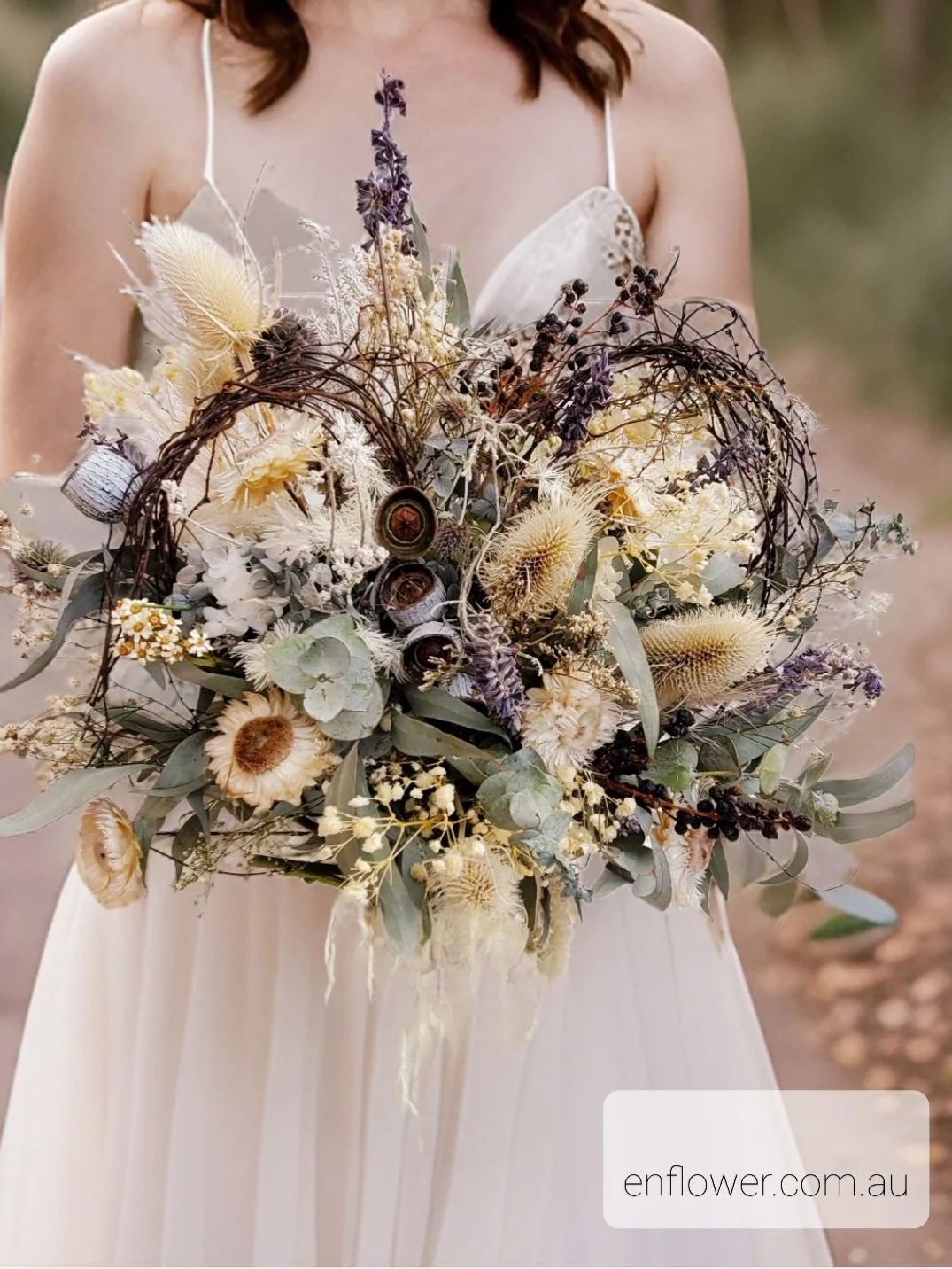 Woman in a white dress holding a large bouquet of dried flowers and foliage, including lavender, eucalyptus, and thistle, in an outdoor setting.