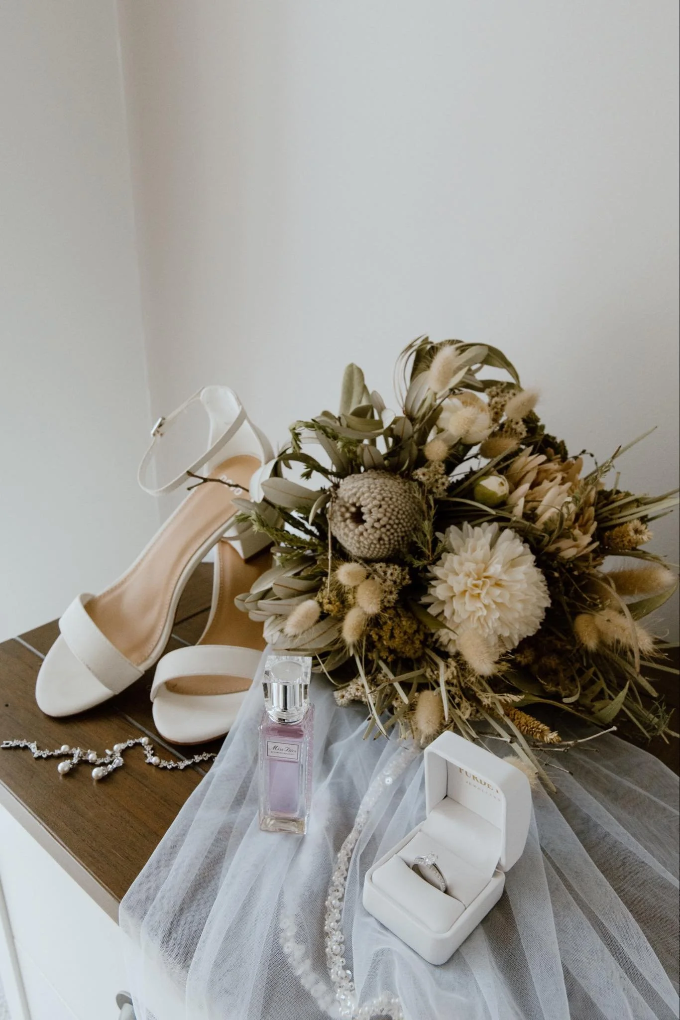 White high-heeled shoes, a perfume bottle, a jewelry box with a ring, a dried flower bouquet, and a pearl necklace displayed on a wooden surface with a sheer cloth.