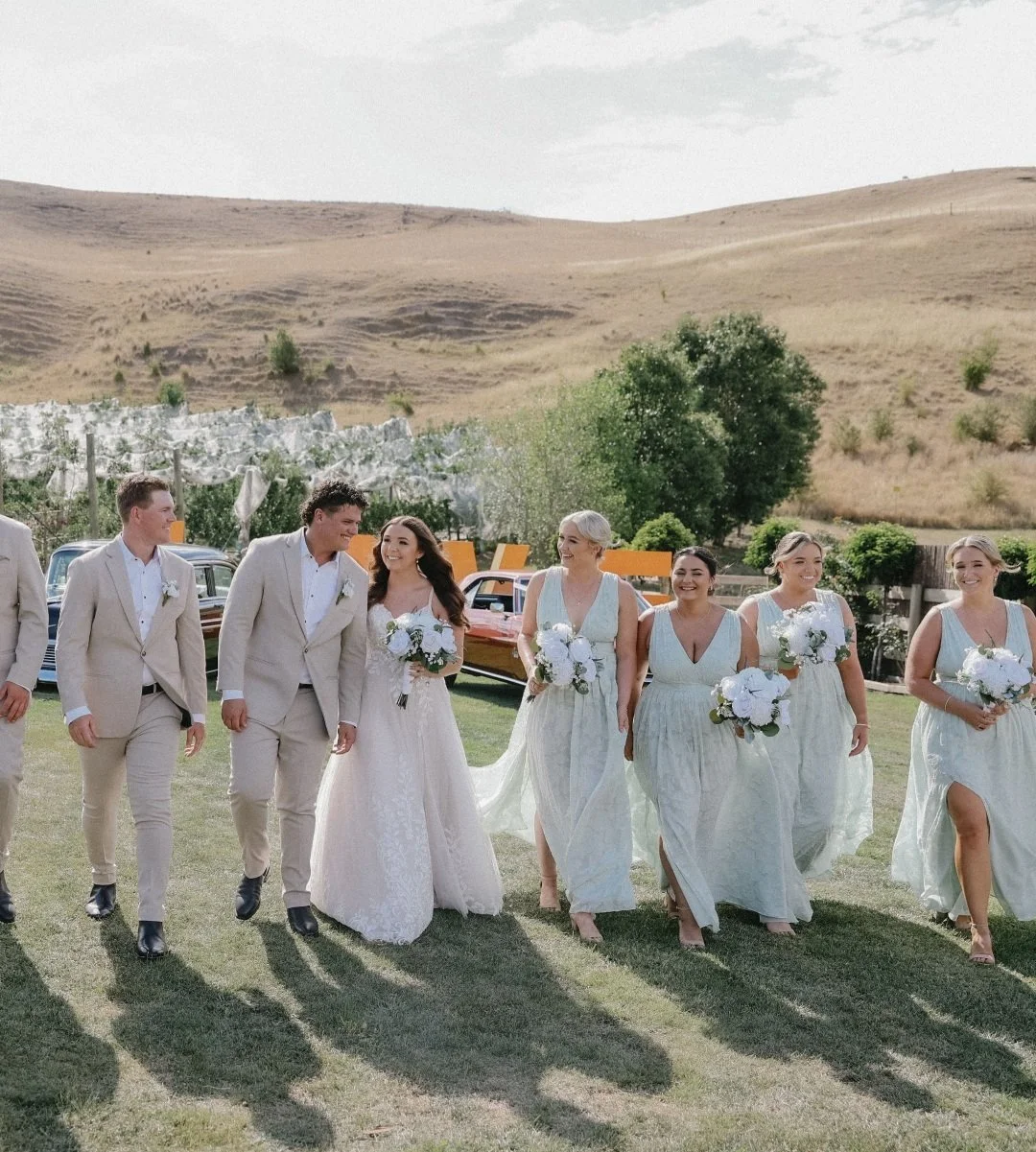 A wedding party walking outdoors on a grassy field with vintage cars and open land in the background. The group consists of a bride in a white wedding dress, groomsmen in beige suits, and bridesmaids in light-colored dresses, all holding white bouque