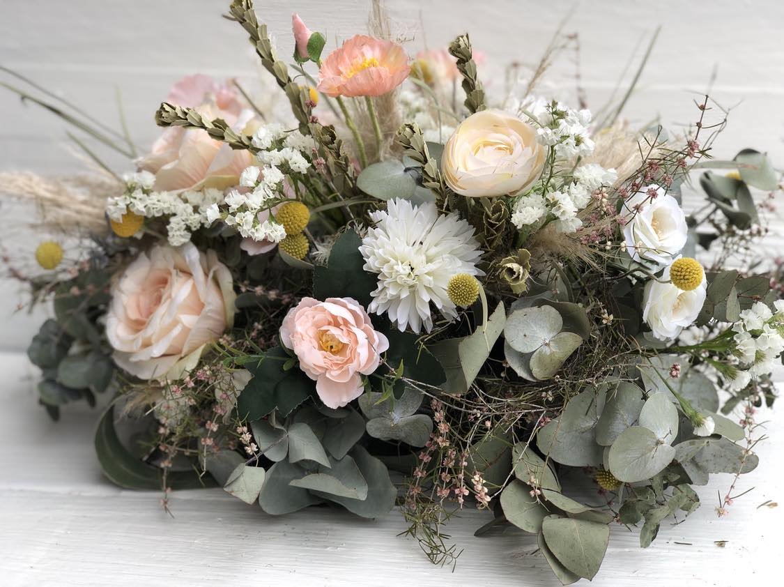 A bouquet of pastel-colored flowers including roses, dahlias, poppies, and small yellow blossoms with greenery, arranged on a light wooden surface.