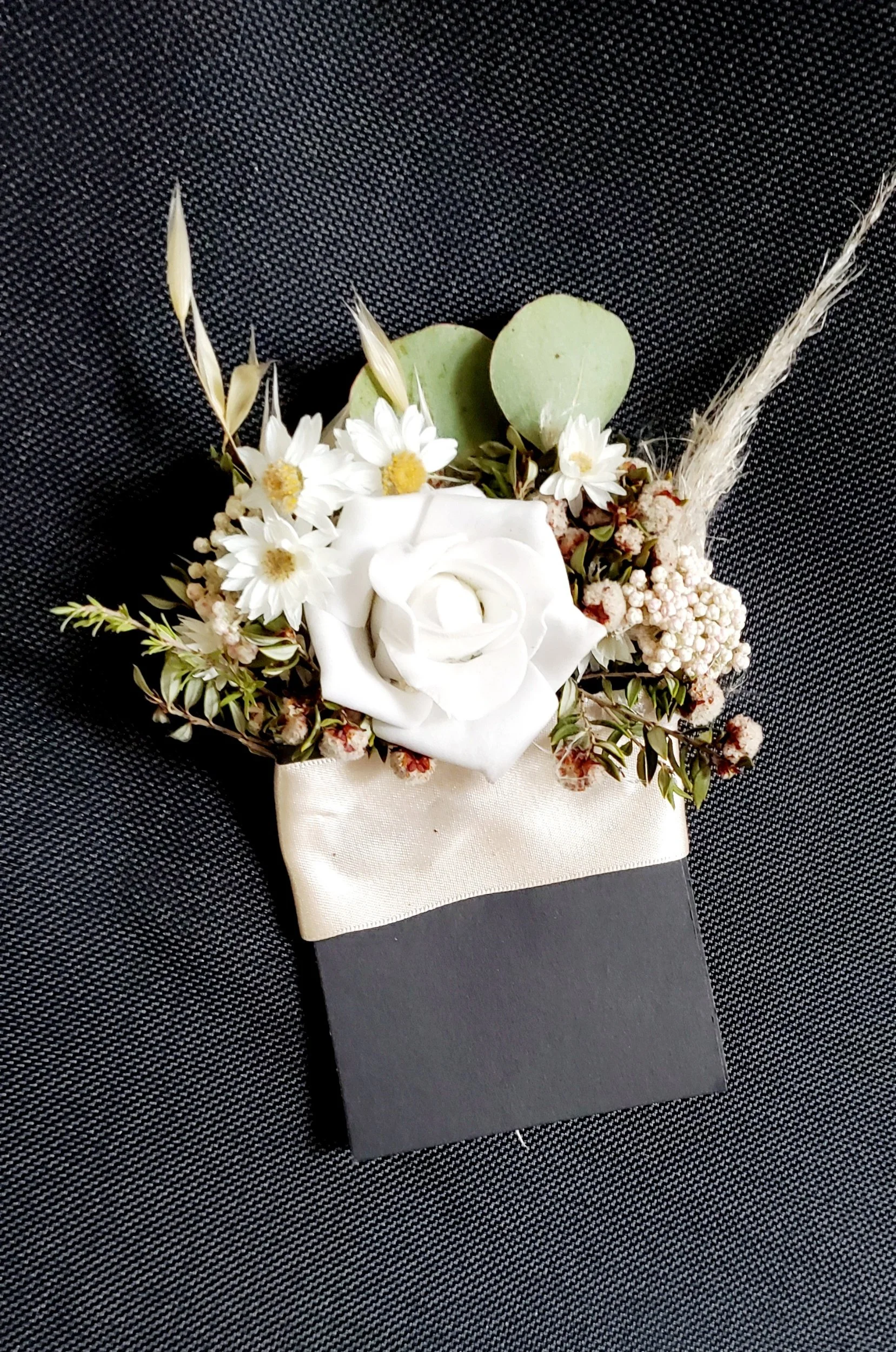 A floral boutonniere with white roses, small white daisies, greenery, and a black and ivory base on a black textured fabric background.