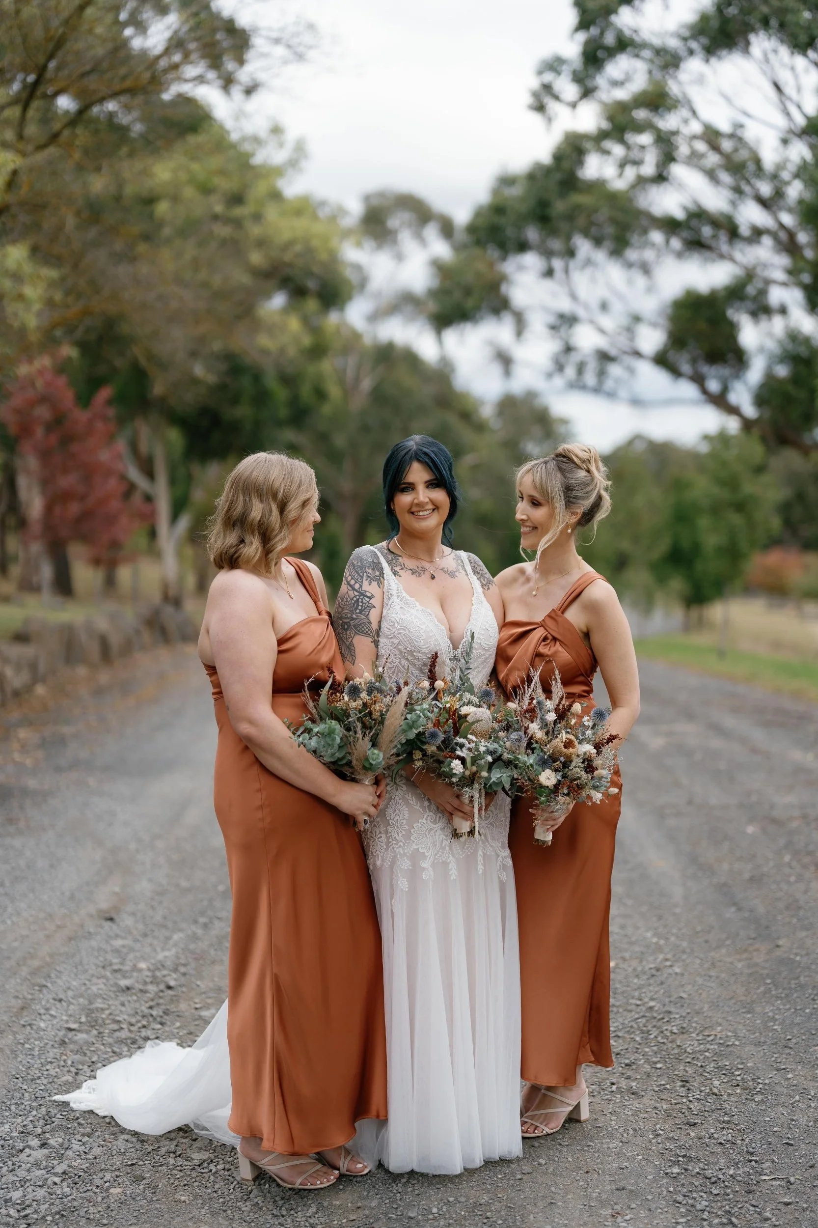 Three women dressed in wedding attire standing outdoors on a gravel path, with trees in the background. One woman is wearing a white wedding dress with lace details, and the other two women are wearing matching bronze-colored bridesmaid dresses. They