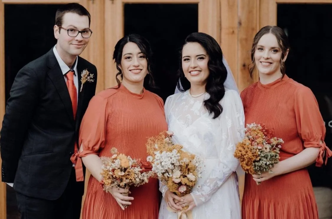 Four people standing together indoors, with two women holding bouquets, one woman in a white dress and veil, likely a bride, and three others in matching orange dresses and formal clothes, possibly at a wedding celebration.