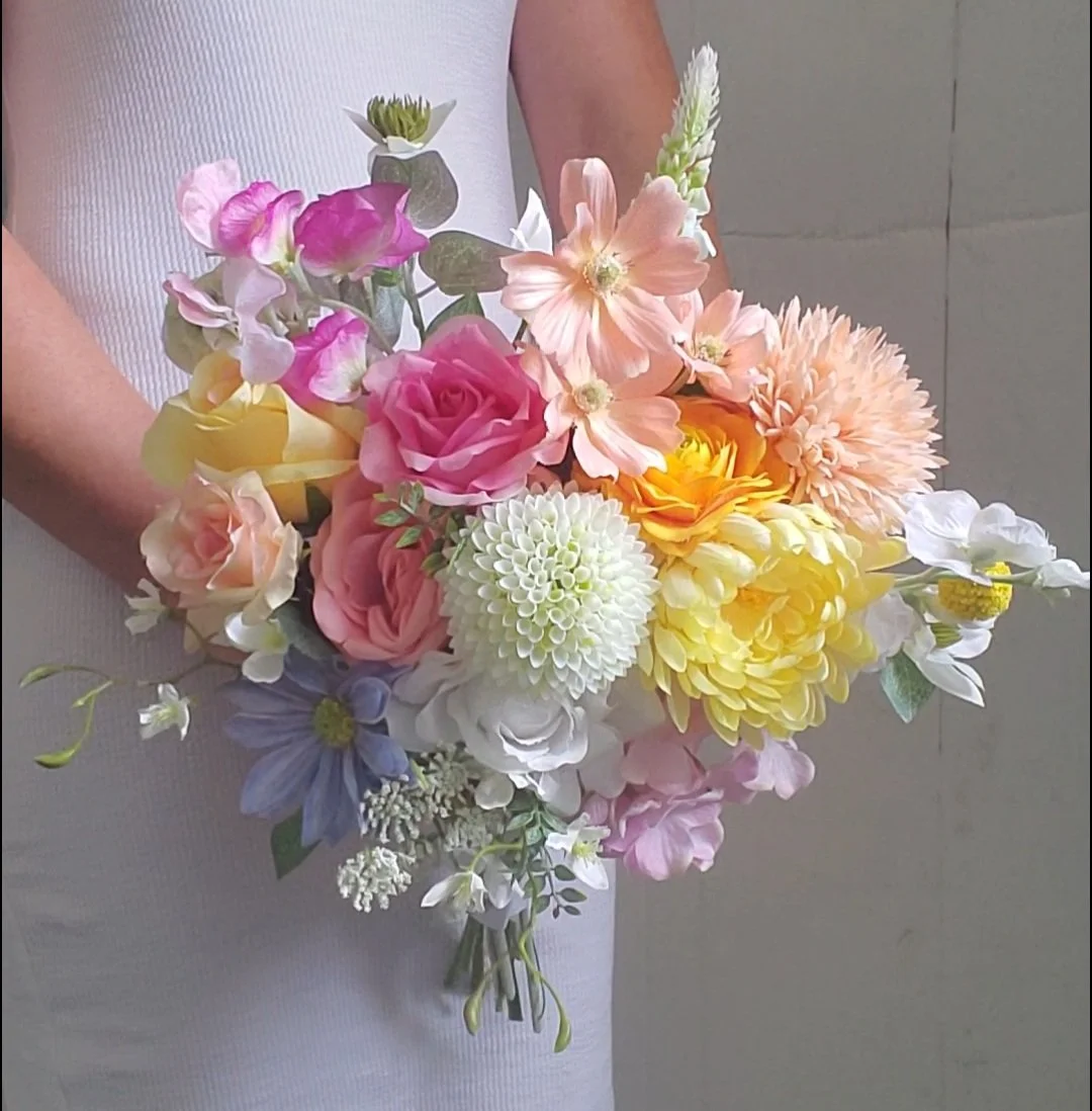 A person holding a colorful bouquet of assorted flowers including roses, dahlias, and other blossoms in shades of pink, yellow, white, and purple.