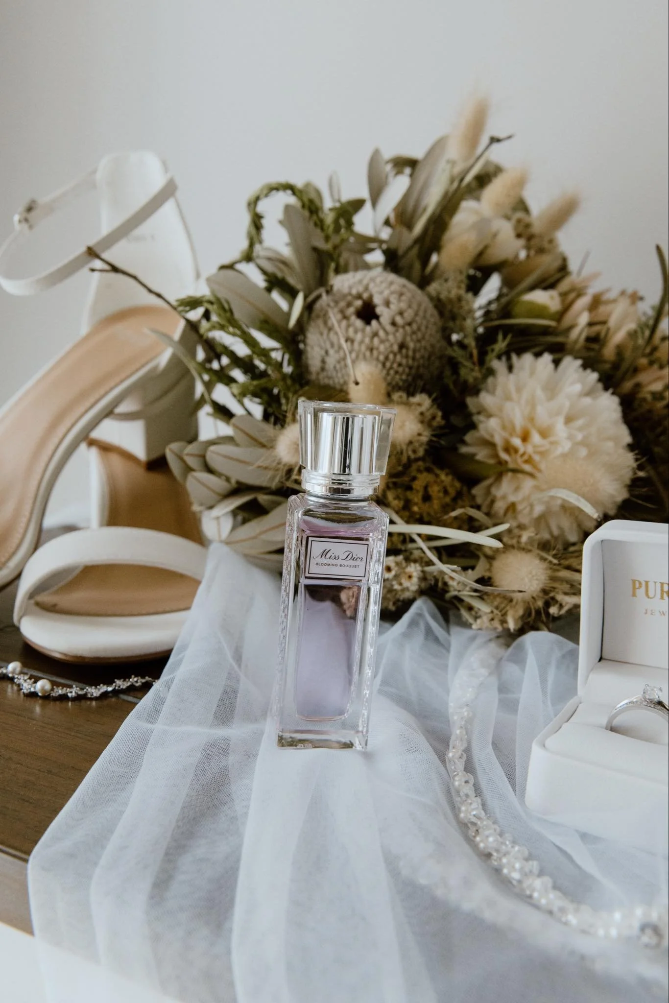A table with a bouquet of dried flowers, a bottle of Miss Dior perfume, a pearl necklace, and a jewelry box with rings. A white sheer fabric is draped on the table.