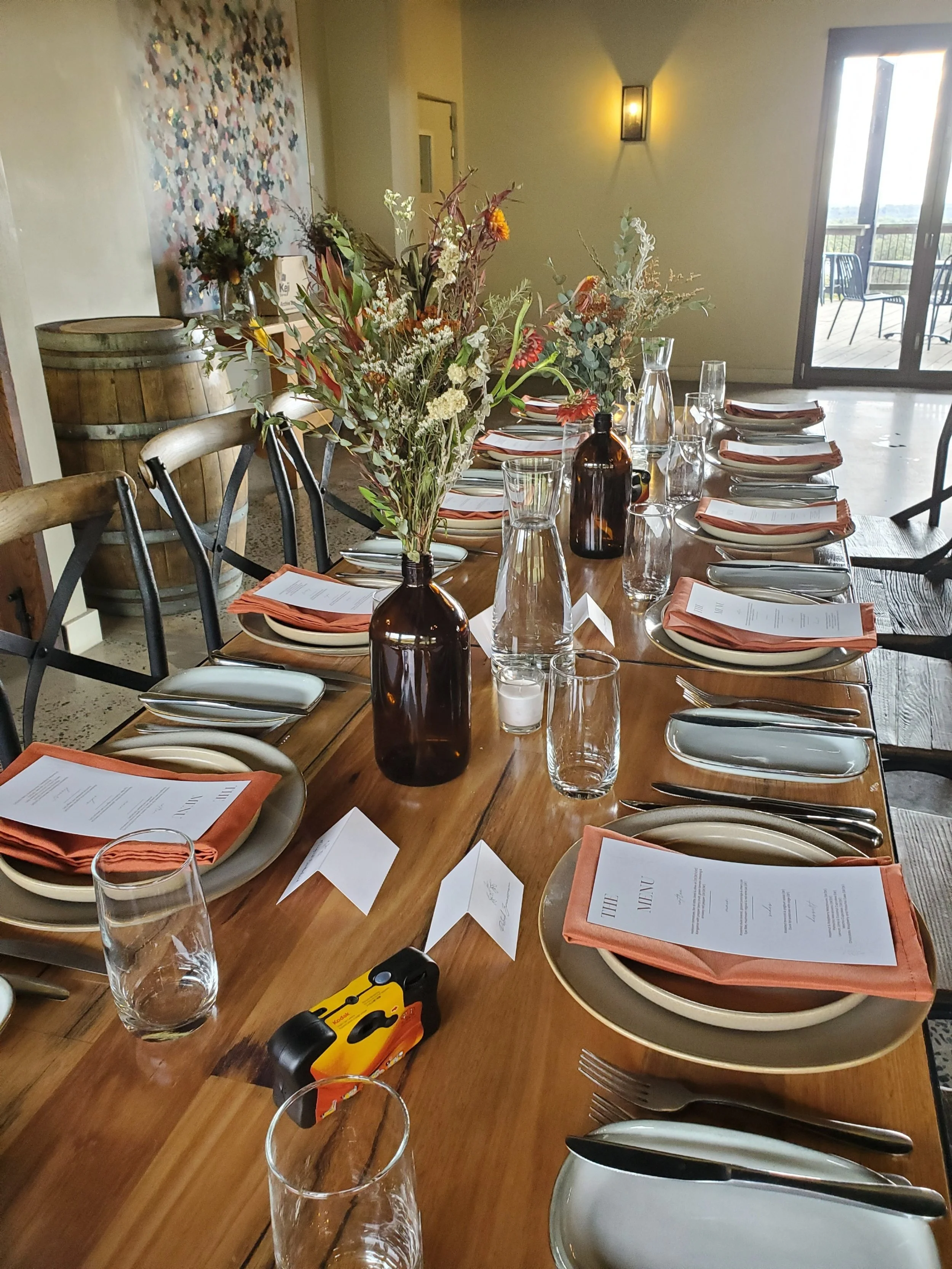 A long wooden dining table set for a meal with plates, utensils, folded napkins, and water glasses, with a centerpiece of tall flowers in brown bottles, in a room with a balcony in the background.