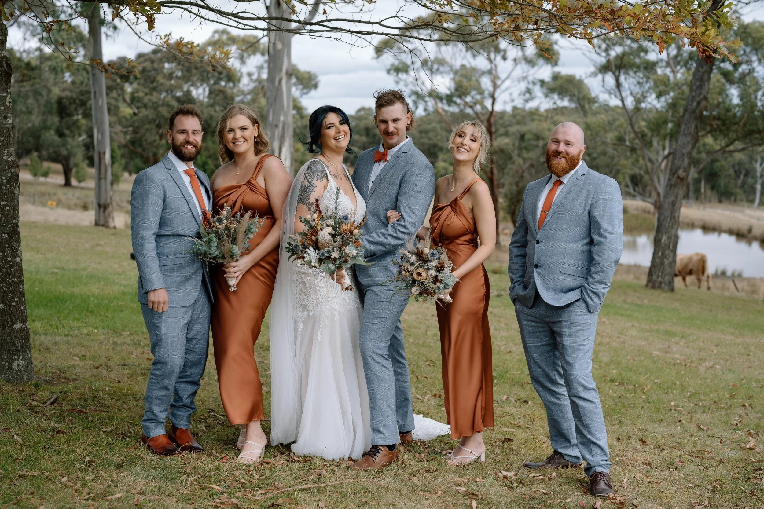 A group of six people dressed in wedding attire, standing outdoors on grass, with trees and a pond in the background. The groom and two bridesmaids hold bouquets, and everyone is smiling.