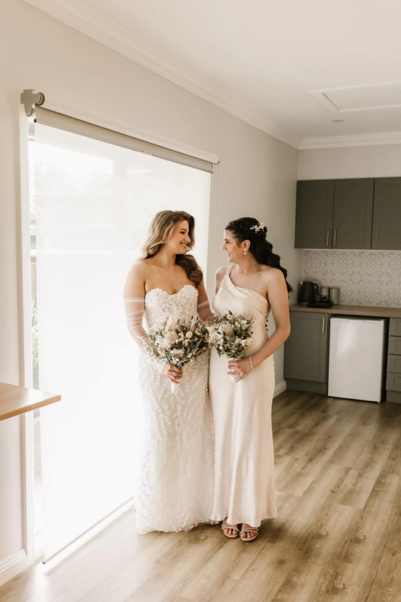 Two women dressed in wedding attire smiling at each other in a bright room, holding bouquets of flowers.