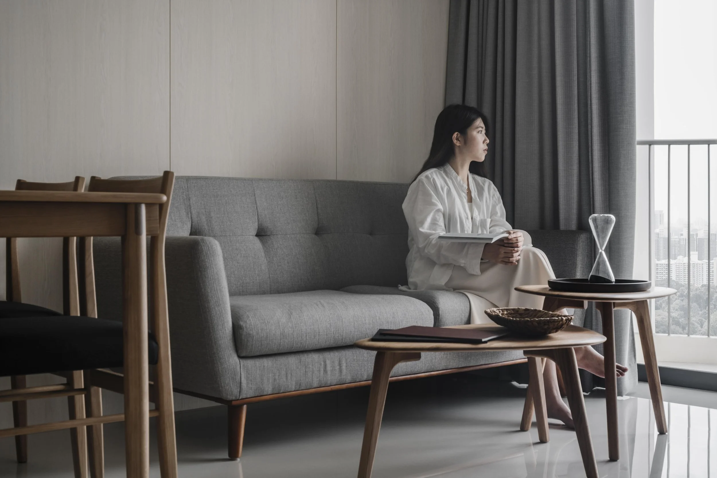 Minimalist Singapore condo living room with grey sofa, wooden furniture and homeowner relaxing by the balcony.