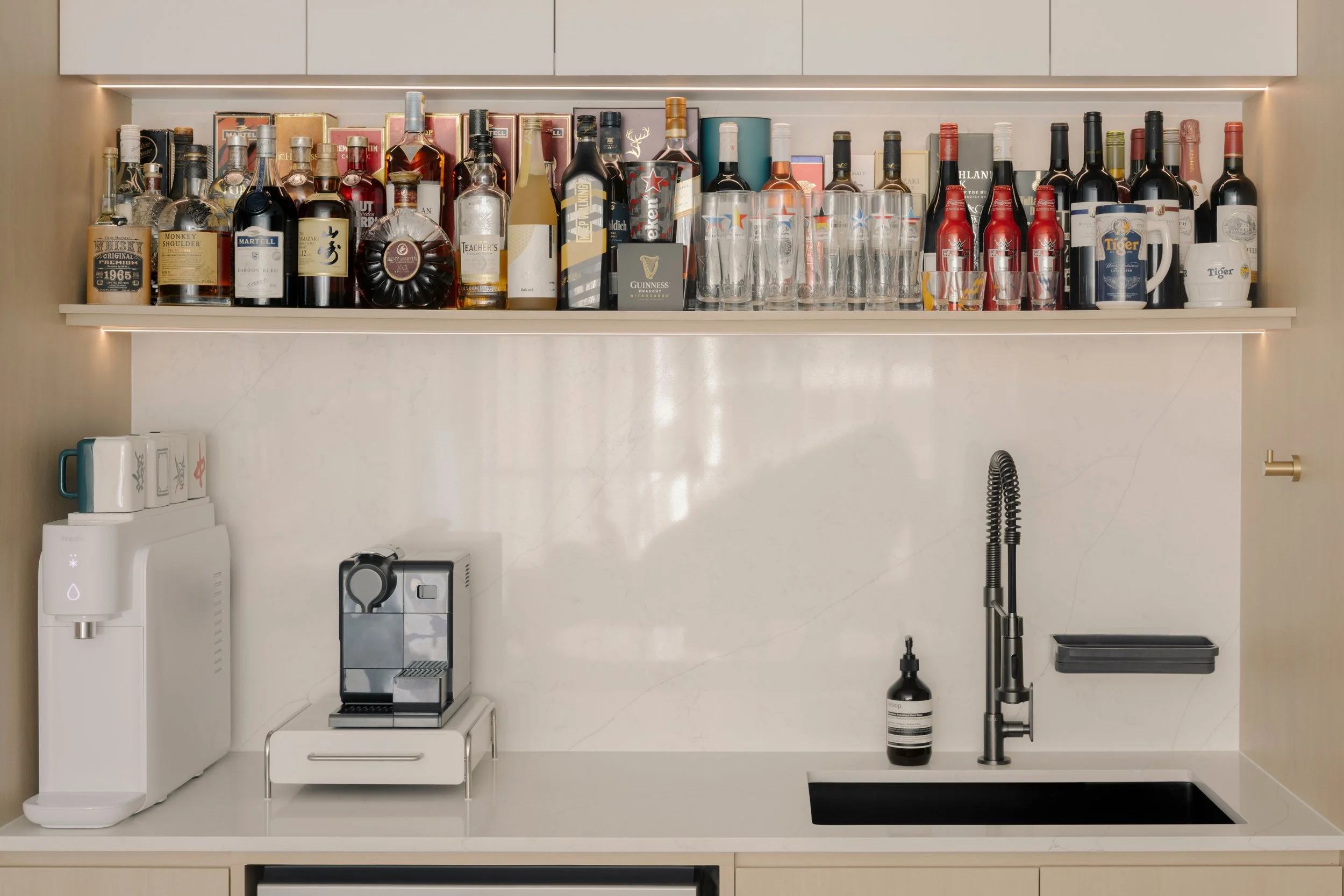Modern home bar with marble backsplash, open display shelf of liquor bottles, built-in coffee machine, and matte black sink in a minimalist interior.