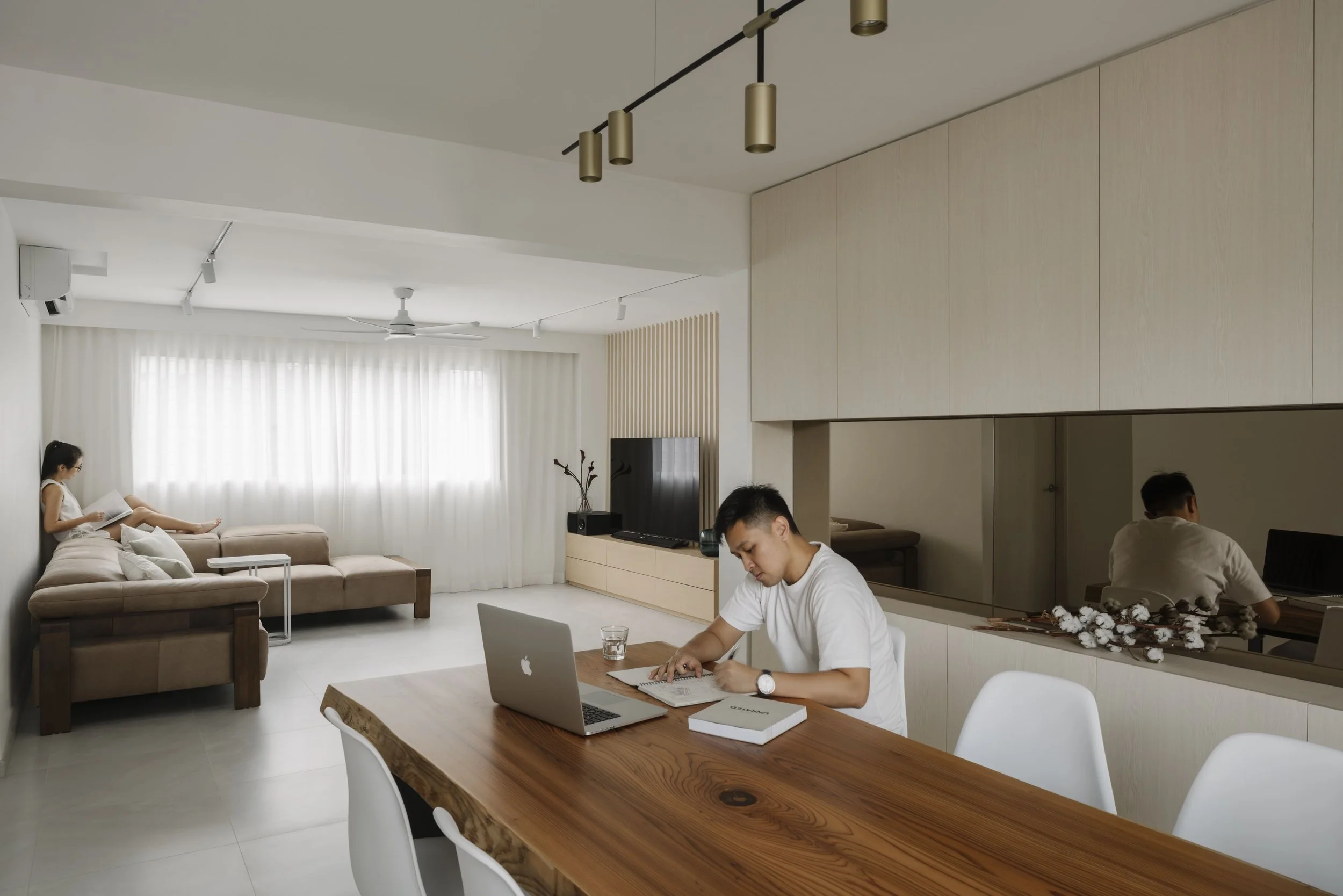Bright minimalist living and dining space with a natural wood dining table, custom built-in cabinetry, and homeowners relaxing in a modern open-concept interior by Lite Design Collaborative in Singapore.