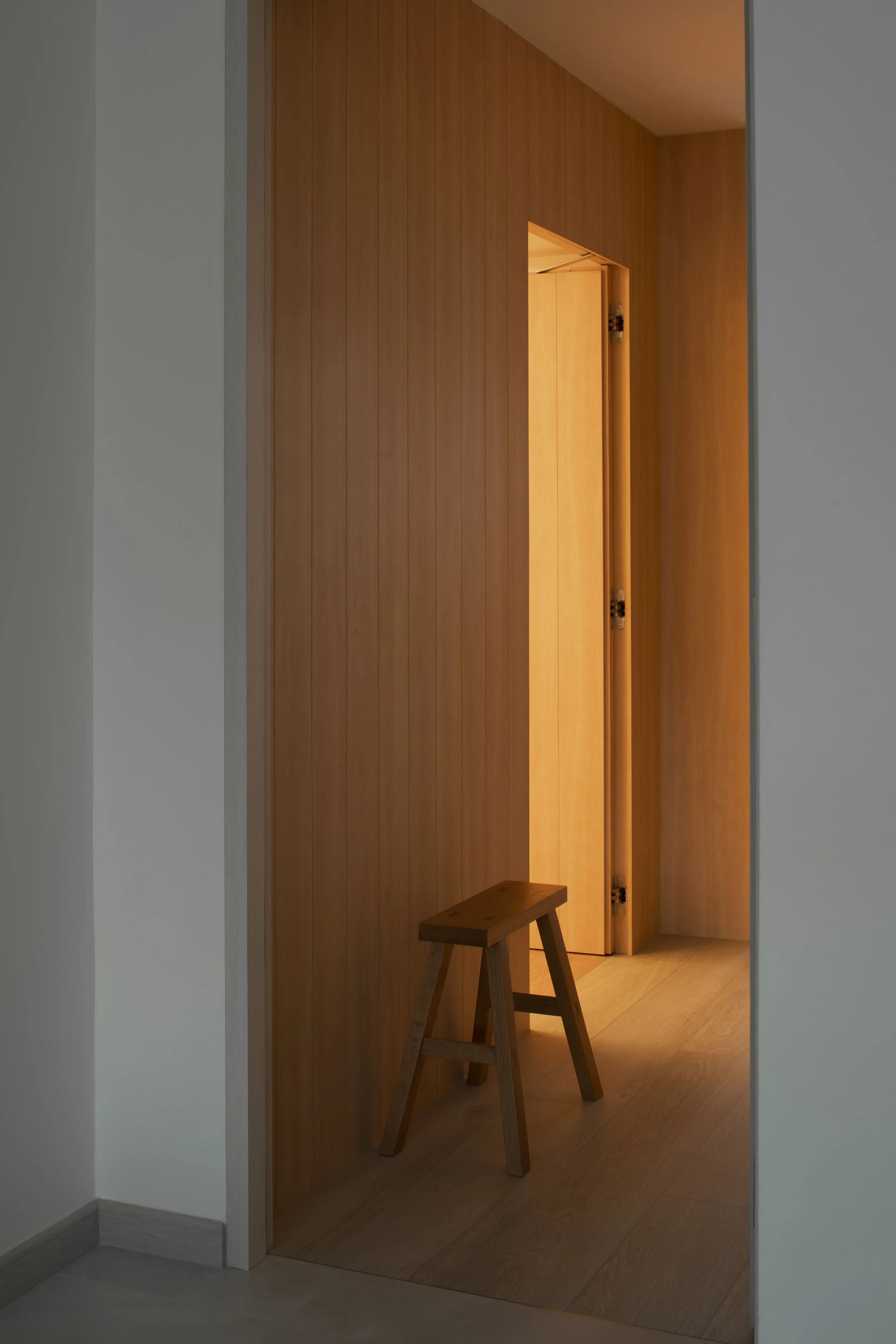 Minimalist hallway with warm wood panel wall, soft ambient lighting, and a simple wooden stool in a modern home interior.
