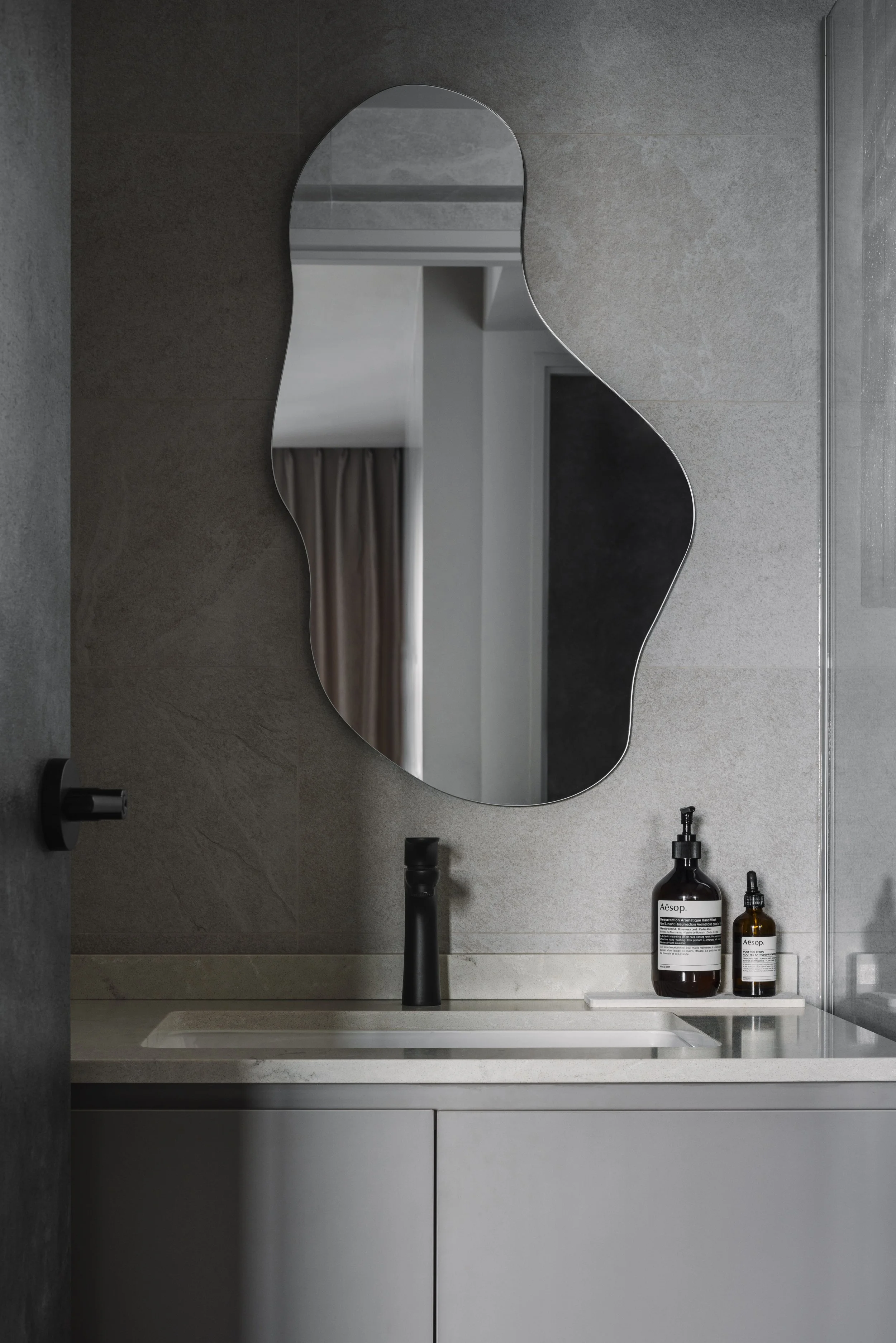 Modern bathroom vanity with an organic-shaped mirror, matte black tap and light grey stone tiles.