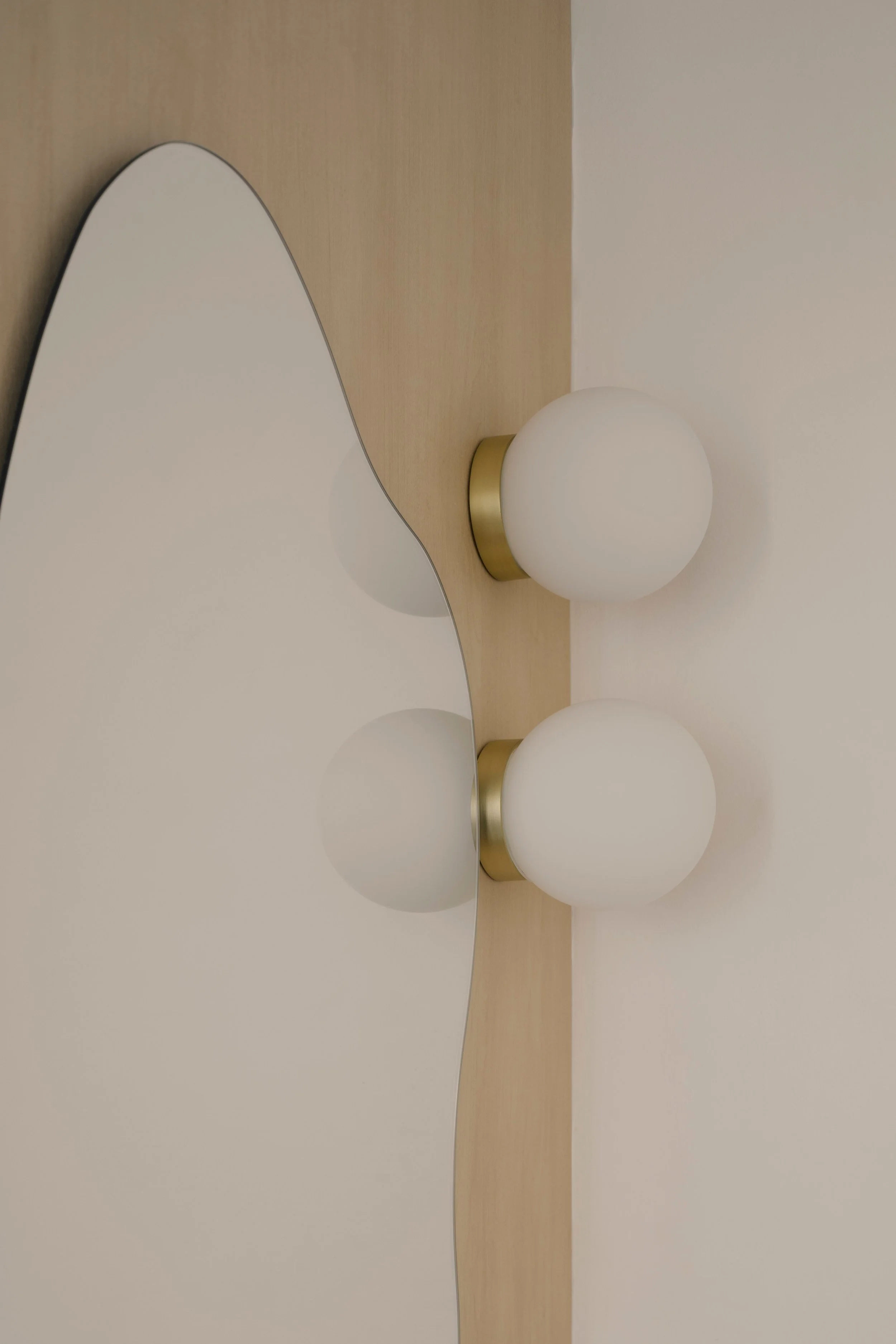 Close-up of organic-shaped mirror with brass double globe wall light against a warm wood panel in a modern minimalist interior.