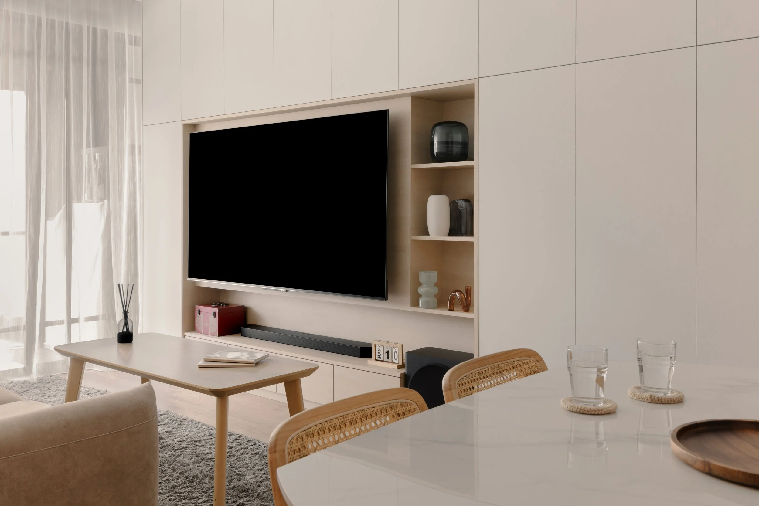 Modern minimalist living room with custom built-in TV feature wall, light-wood shelving, and a bright open dining area.