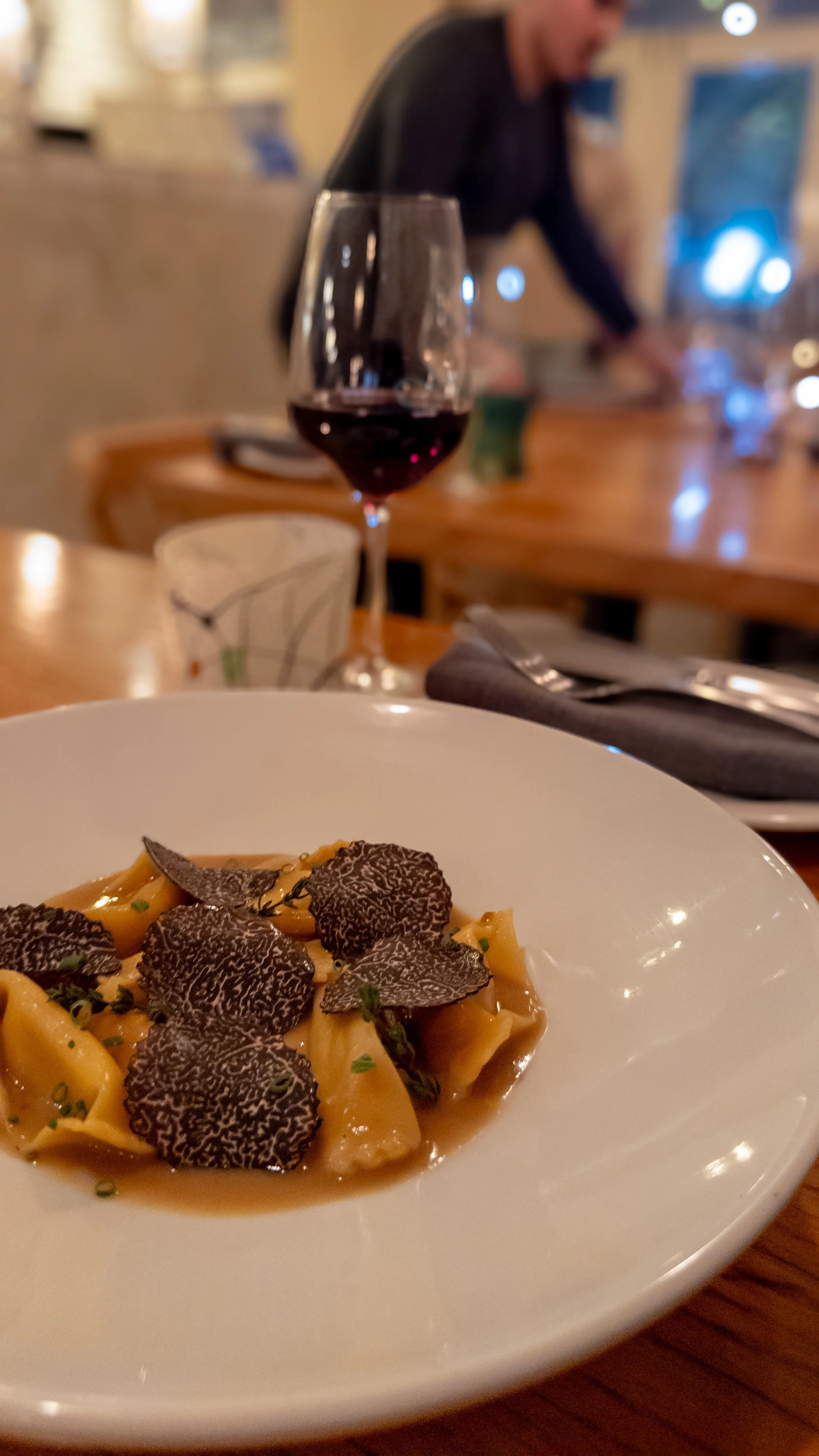 A white plate with pasta topped with black truffle slices on a wooden table, with a glass of red wine and a glass of water in the background, in a dimly lit restaurant.