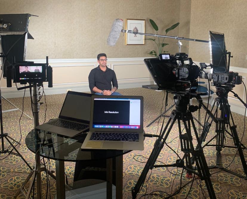 Behind-the-scenes of a video interview setup with a person seated, surrounded by professional camera and lighting equipment, and a laptop on a table displaying "Info Resolution".