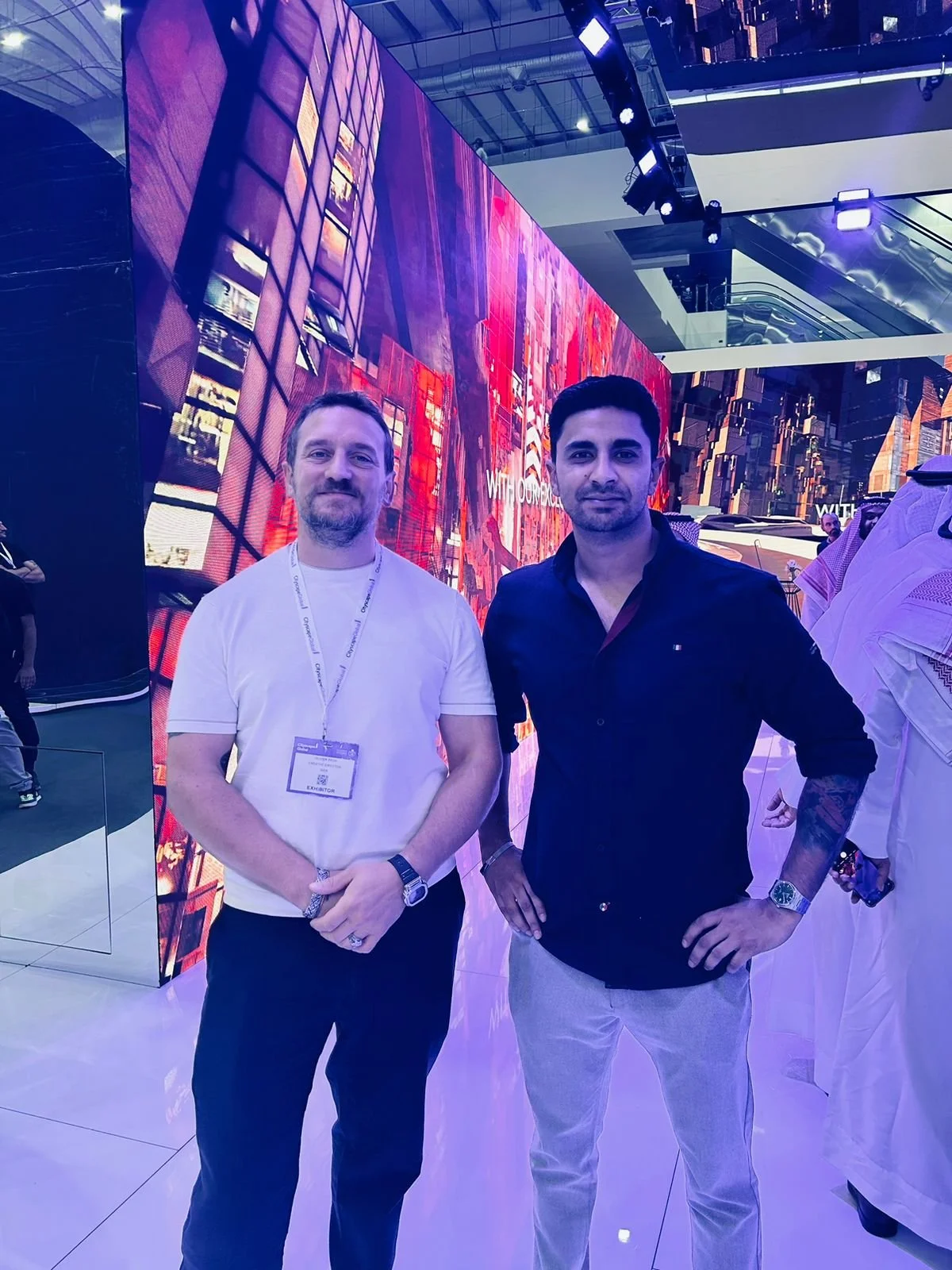 Two men posing indoors in front of a vibrant digital display, featuring cityscape imagery. The man on the left is wearing a white shirt with a name tag, and the man on the right is in a dark shirt with tattoos on his arm. Other people are visible in the background.