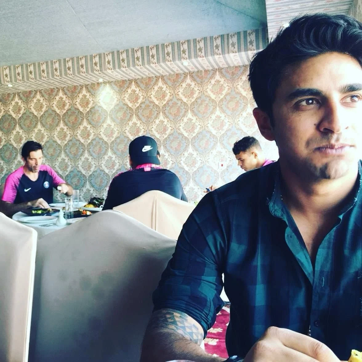 Man in a patterned shirt sitting in a restaurant with a decorative wallpaper and two other people in the background eating.