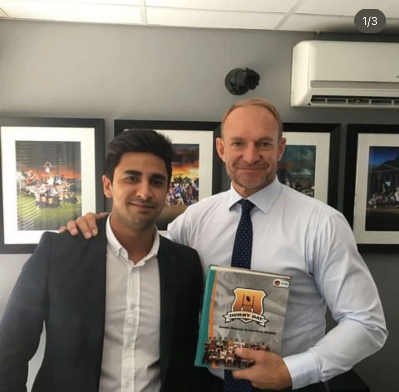 Two men in business attire standing in front of framed pictures, one holding a book titled "Derby Day."