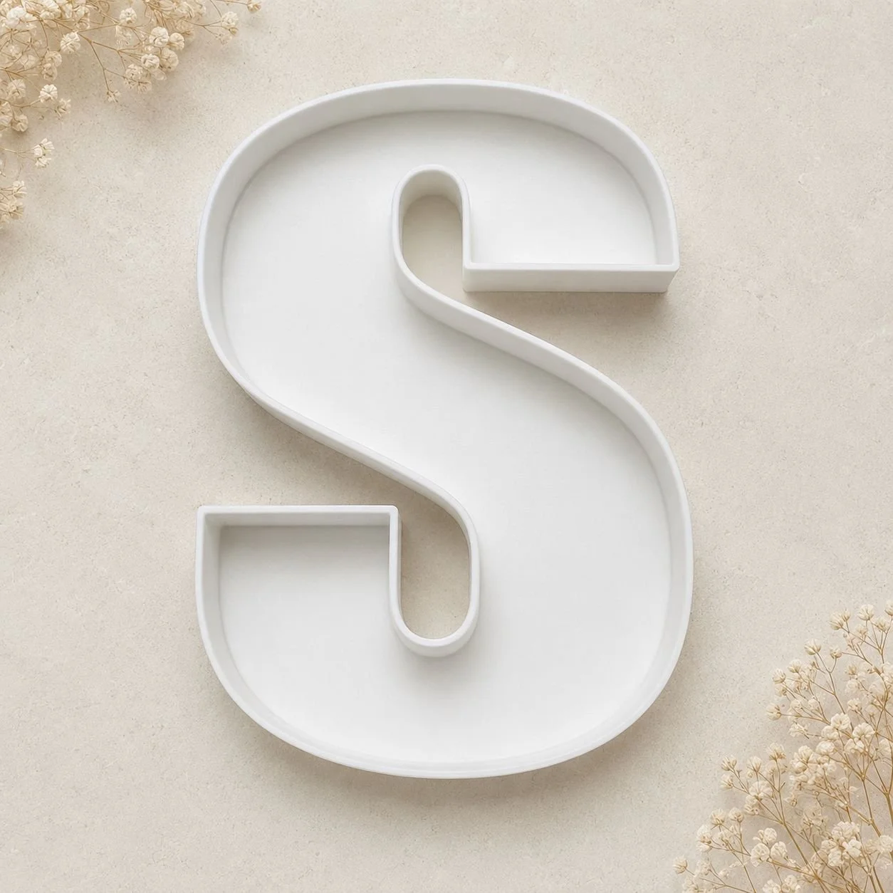 A white, S-shaped decorative dish with a small rectangular section at the bottom, placed on a neutral-colored surface with some dried flowers in the corners.