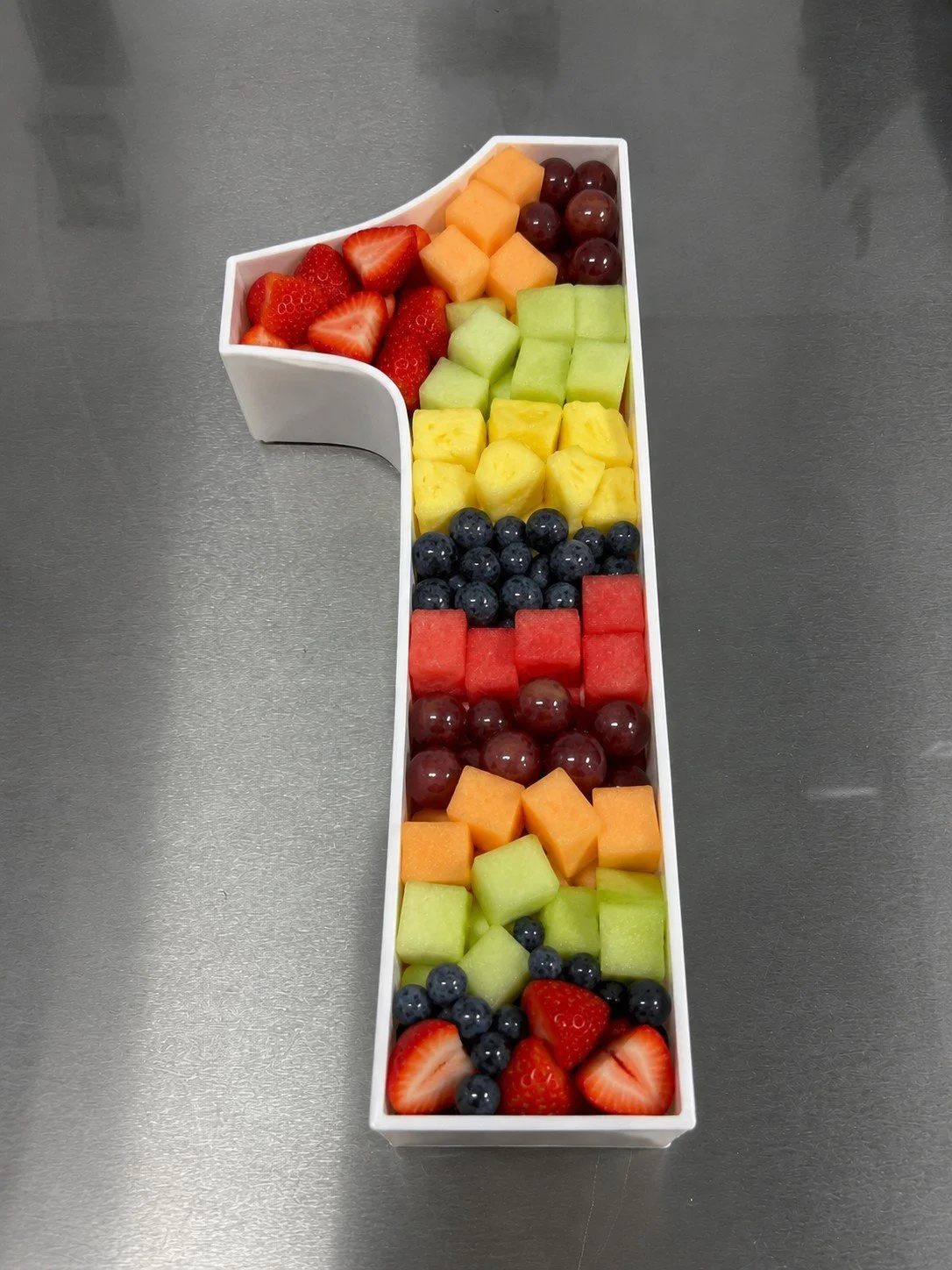 A white number 1 shaped container filled with assorted cut fresh fruits, including strawberries, grapes, honeydew melon, pineapple, blueberries, watermelon, cantaloupe, and cantaloupe, arranged in sections on a glass surface.