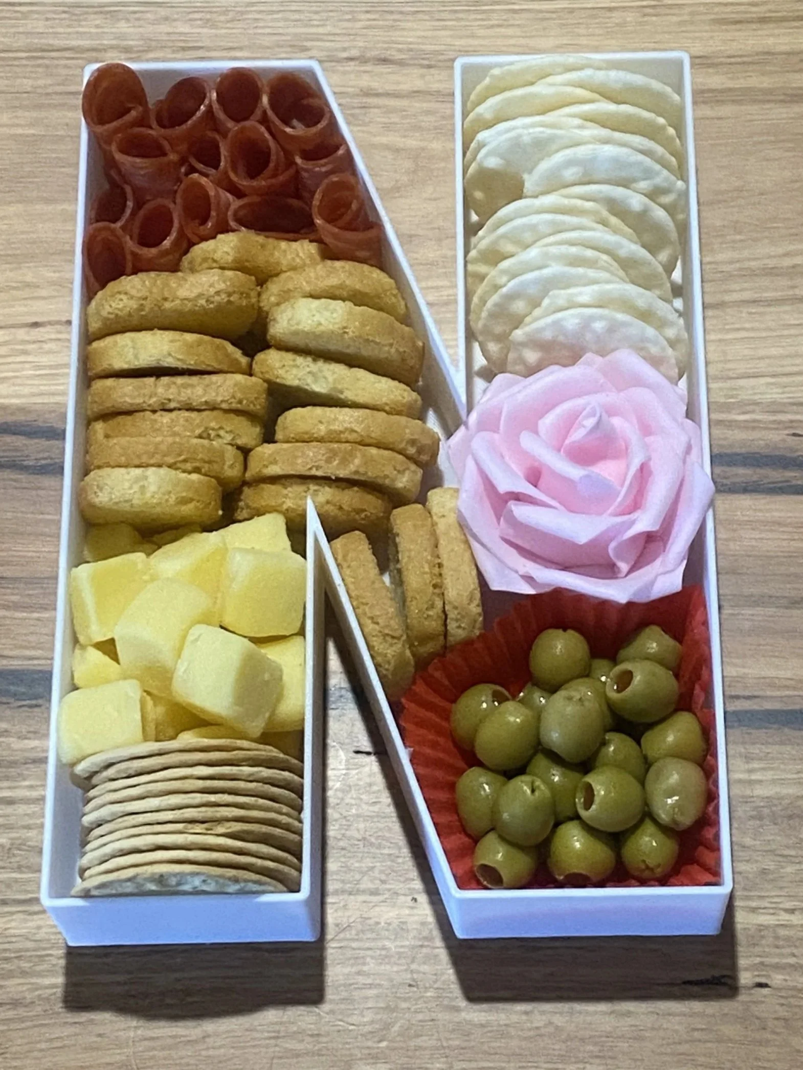 A white rectangular platter with separated compartments filled with various snacks: cheese cubes, crackers, potato chips, slices of salami, chicken nuggets, green olives in a red paper cup, and a pink artificial rose.