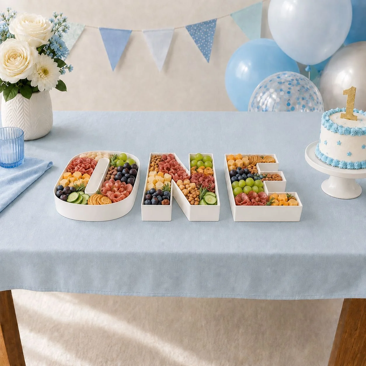 Table decorated for a child's first birthday with the spelled out word "one" in individual white fillable letter boxes with fruit and charcuterie snacks, a blue and white cake with a gold number one topper, and blue and white balloons.
