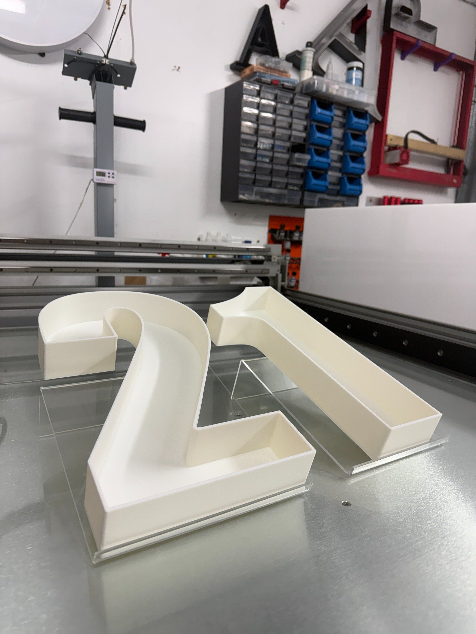 A workshop or manufacturing space with a white 3D letters sculpture on a gray work surface, electrical components, tools, storage drawers, and a red frame on the wall.