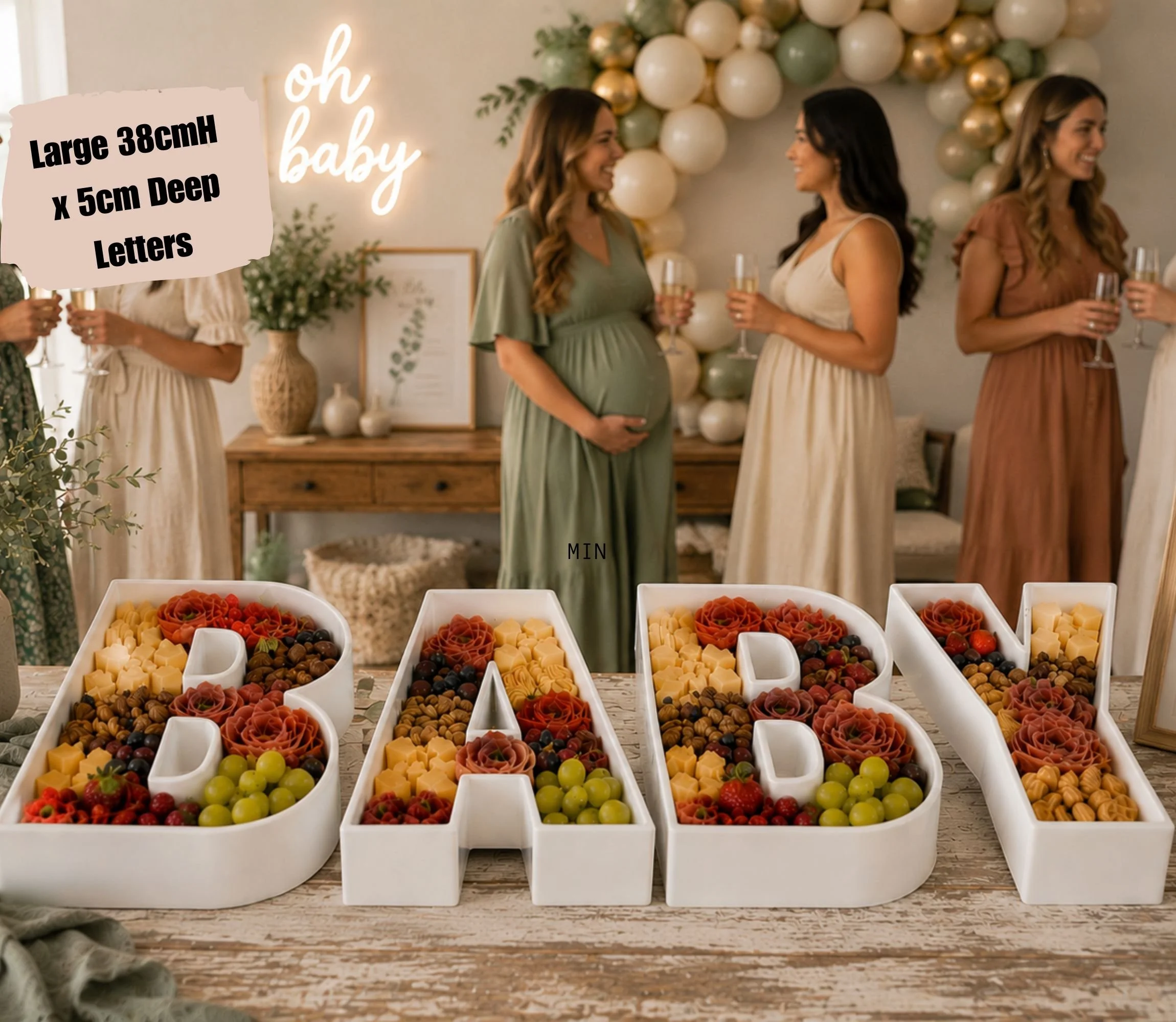 Baby shower letters setup with a table of colorful candies in white containers, a balloon garland, and a neon sign that says "oh baby". Three women, including a pregnant woman, are holding glasses and talking.