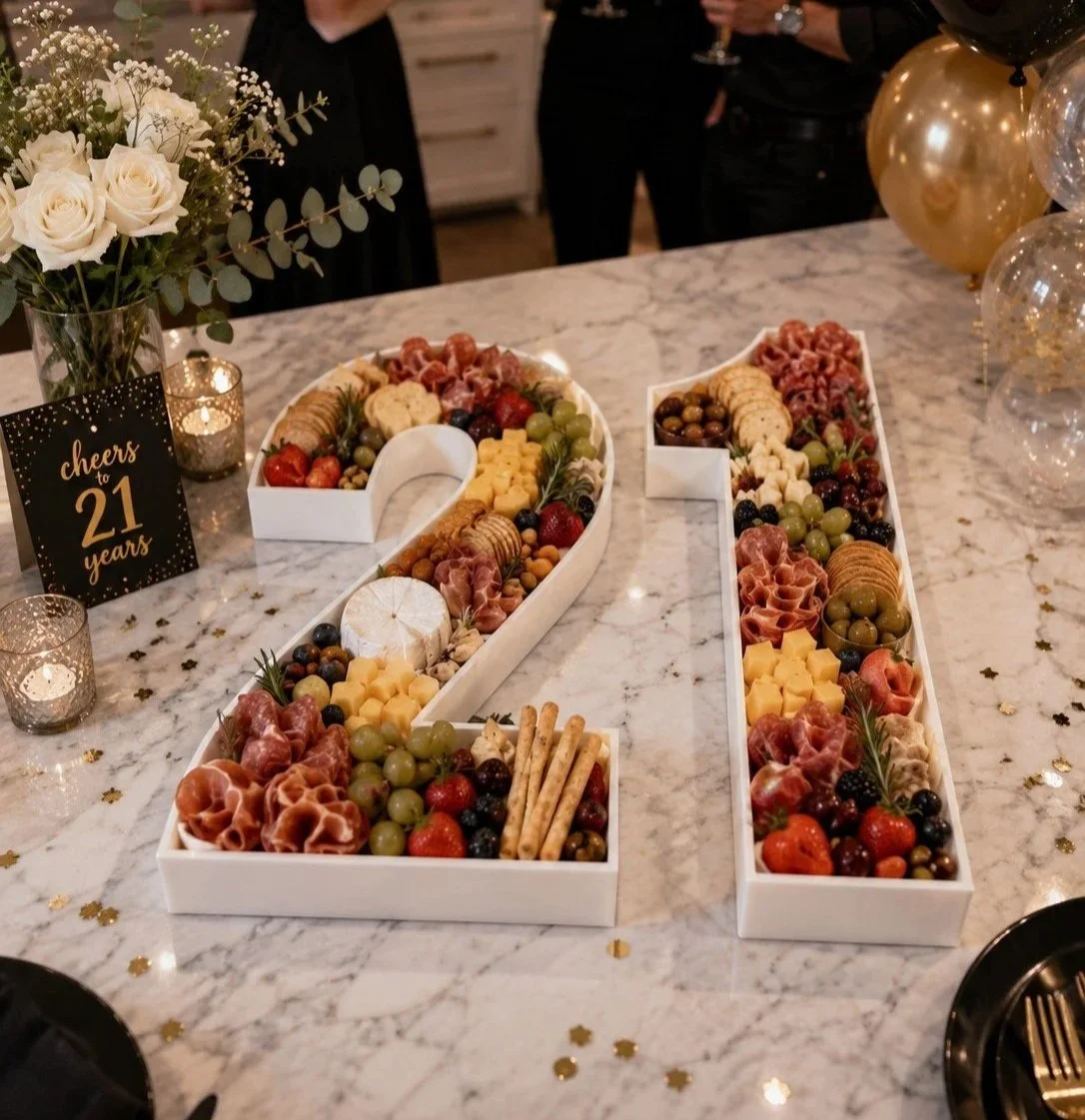 Large decorative platter numbers '21' filled with assorted cheeses, meats, grapes, strawberries, and crackers on a marble table at a celebration.