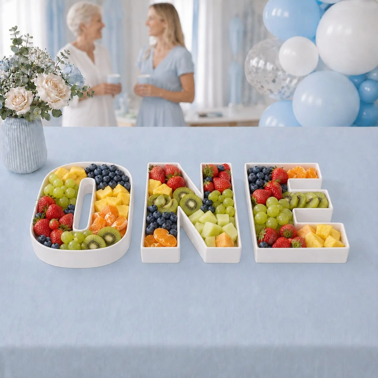 Decorative white fillable box letters spelling 'ONE' filled with assorted fresh fruits, on a light blue tablecloth at a celebration.