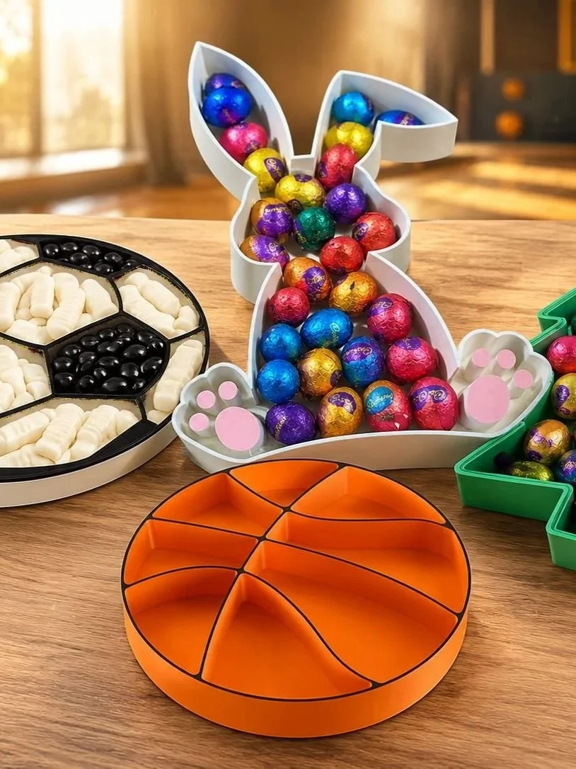 Colorful foil-wrapped chocolates in a bunny-shaped tray, a division of candy on an orange hockey puck-shaped container, and a round divided tray with white and black candies on a wooden table.