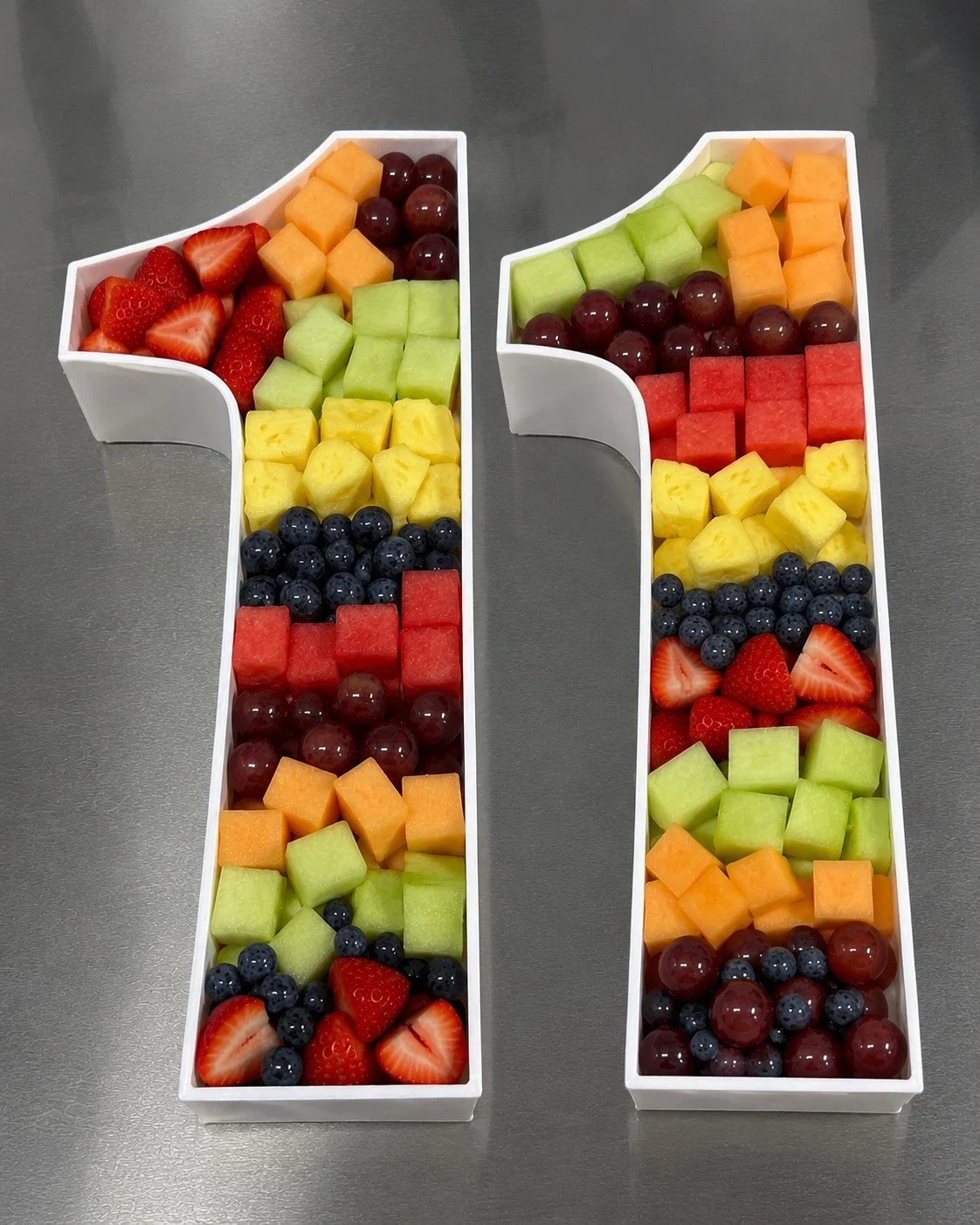 Two large white platter number trays filled with assorted cut fresh fruits including strawberries, cantaloupe, honeydew, watermelon, pineapple, blueberries, and red grapes, arranged in a decorative pattern.