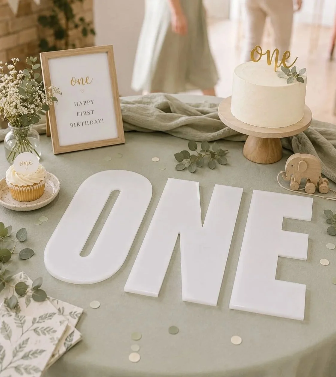 First birthday celebration table with a white cake, an acrylic letter display spelling 'one', a framed sign reading 'Happy First Birthday!', a small wooden elephant toy, a vase with flowers, and large white letter tiles spelling one