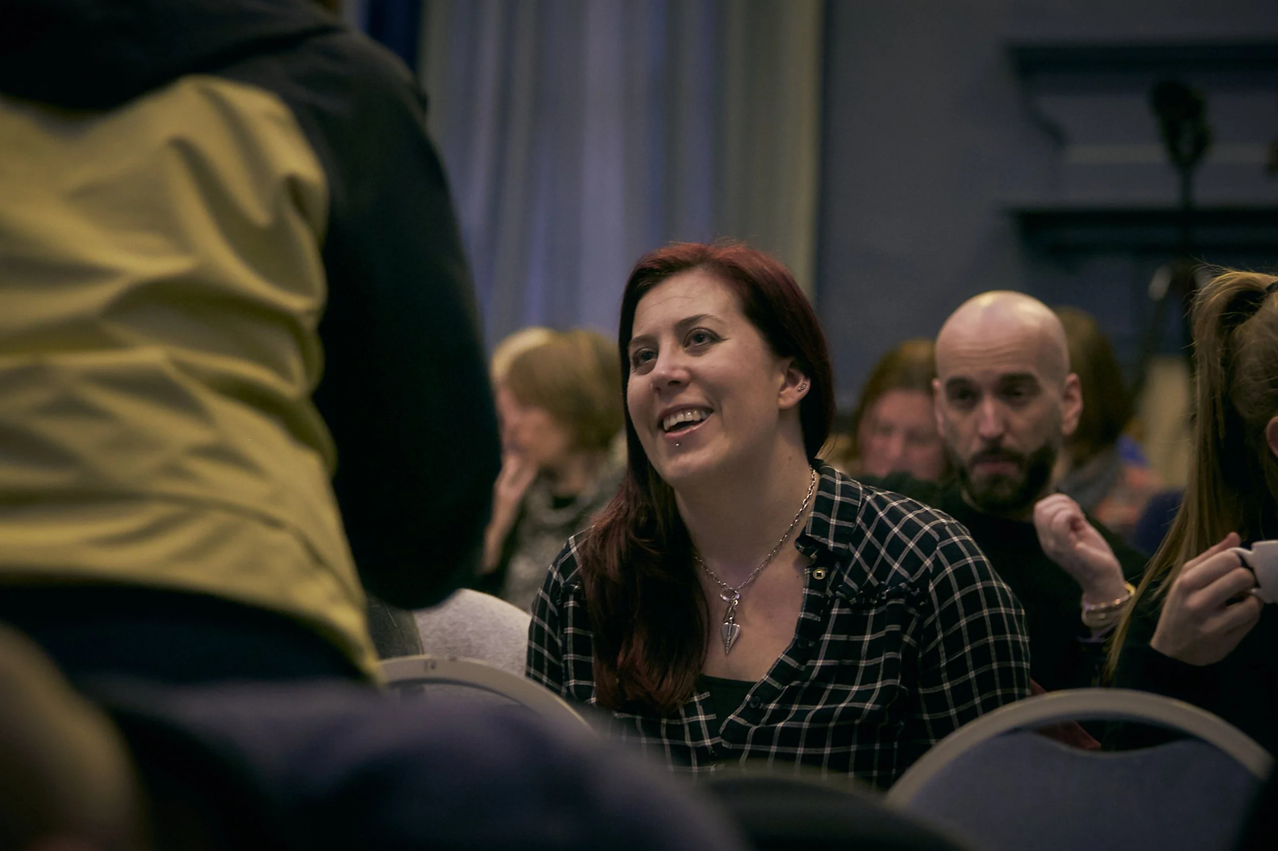 A woman with red hair smiling, sat in an audience. She is watching a presentation.