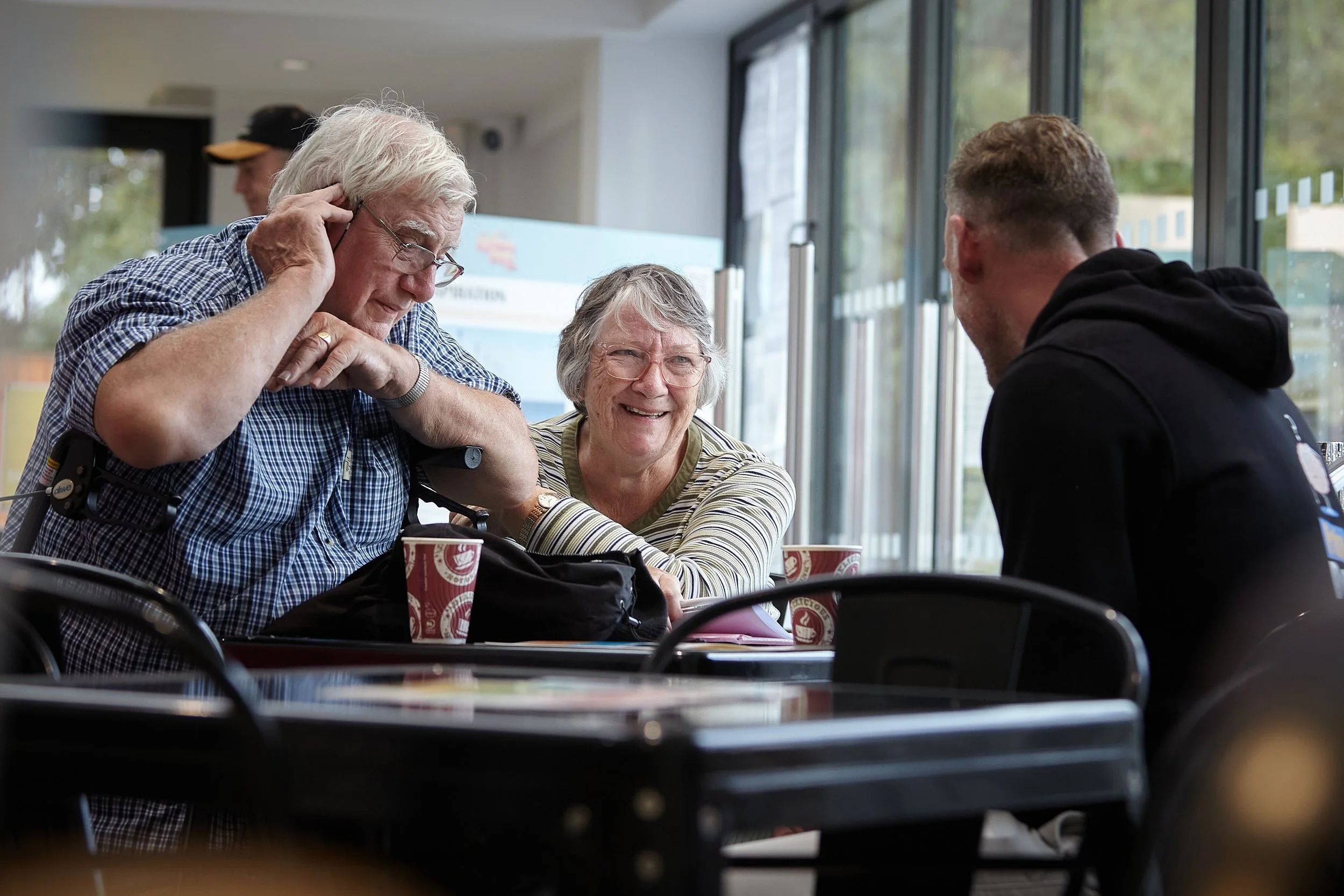 A Flippin' Pain staff member with his back to the camera, talking to an elderly couple over cups of coffee.