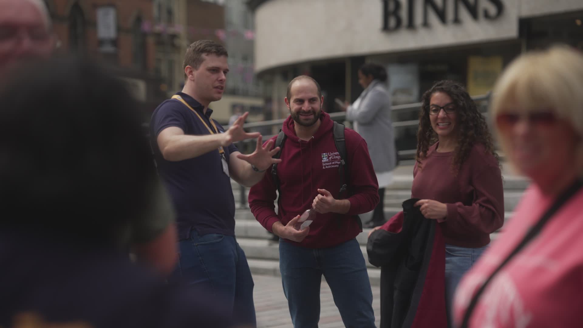 A group of five people standing outdoors in a conversation, with three in focus. One man is gesturing with his hands, smiling, and wearing a maroon hoodie with a backpack. Another man appears to be listening with a neutral expression, wearing a navy blue shirt. A woman with curly hair, glasses, and a maroon top is smiling. Two women are partially visible in the foreground, one with blonde hair and the other with darker hair and glasses, with blurred faces.