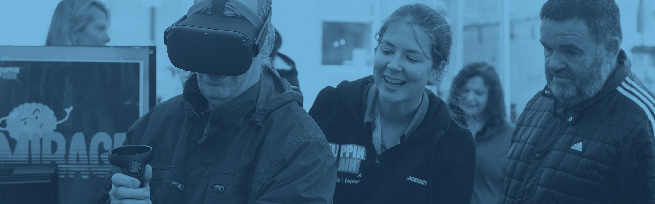 A photograph of a man wearing a VR headset. Behind him is a woman trying to guide him.