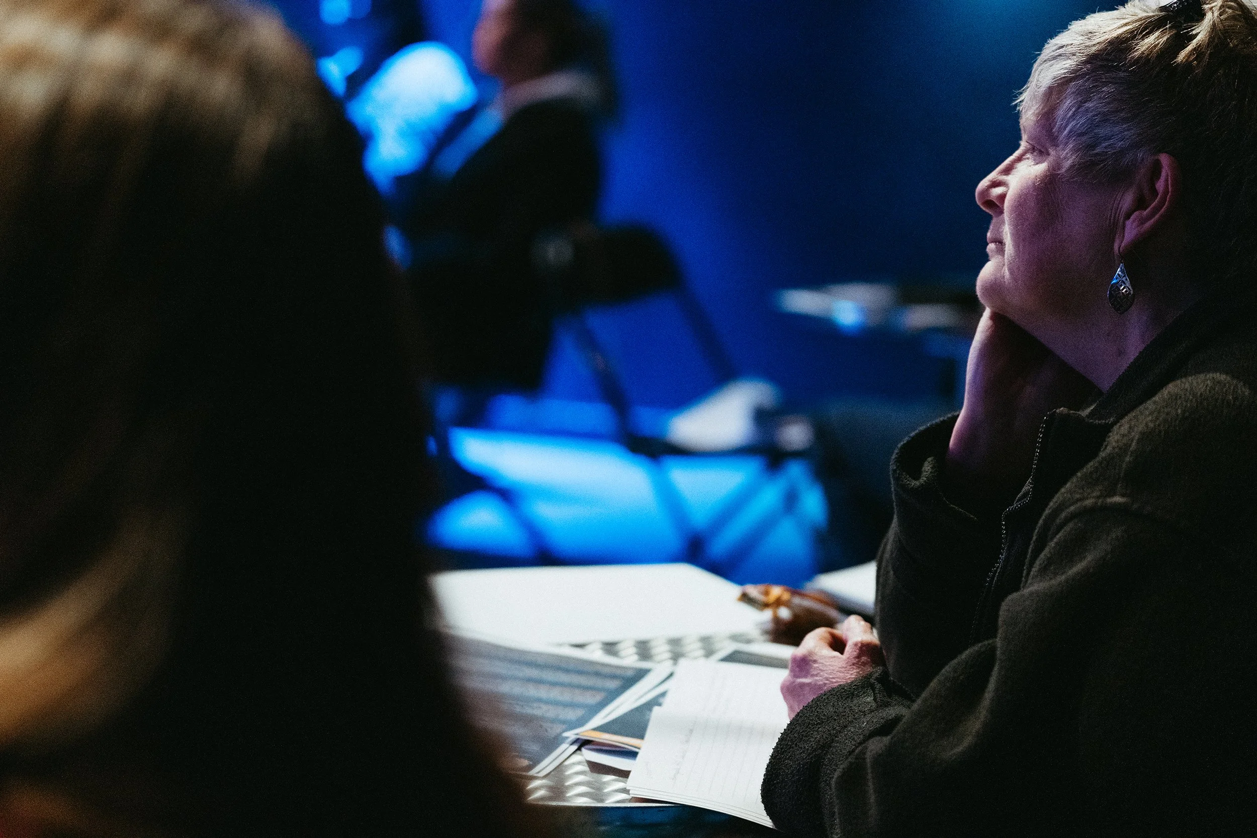 A photograph of a woman sat at a table at an event, watching and smiling.