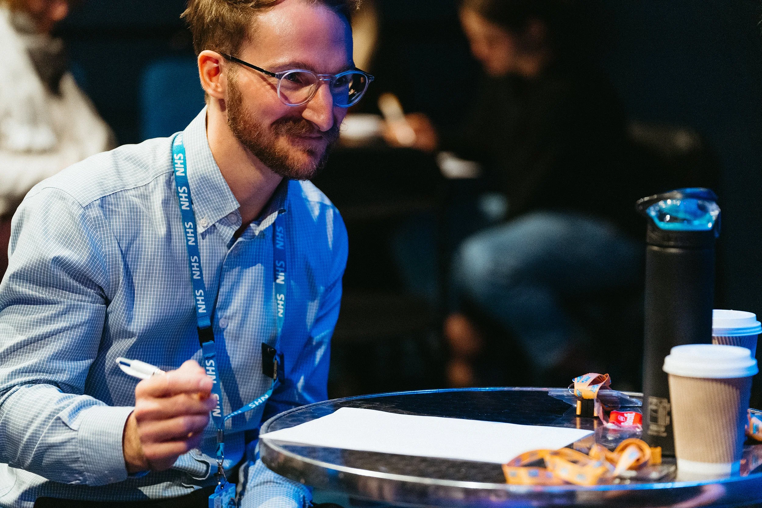 An NHS employee smiling. He is sat at a table with a pen in his hand.