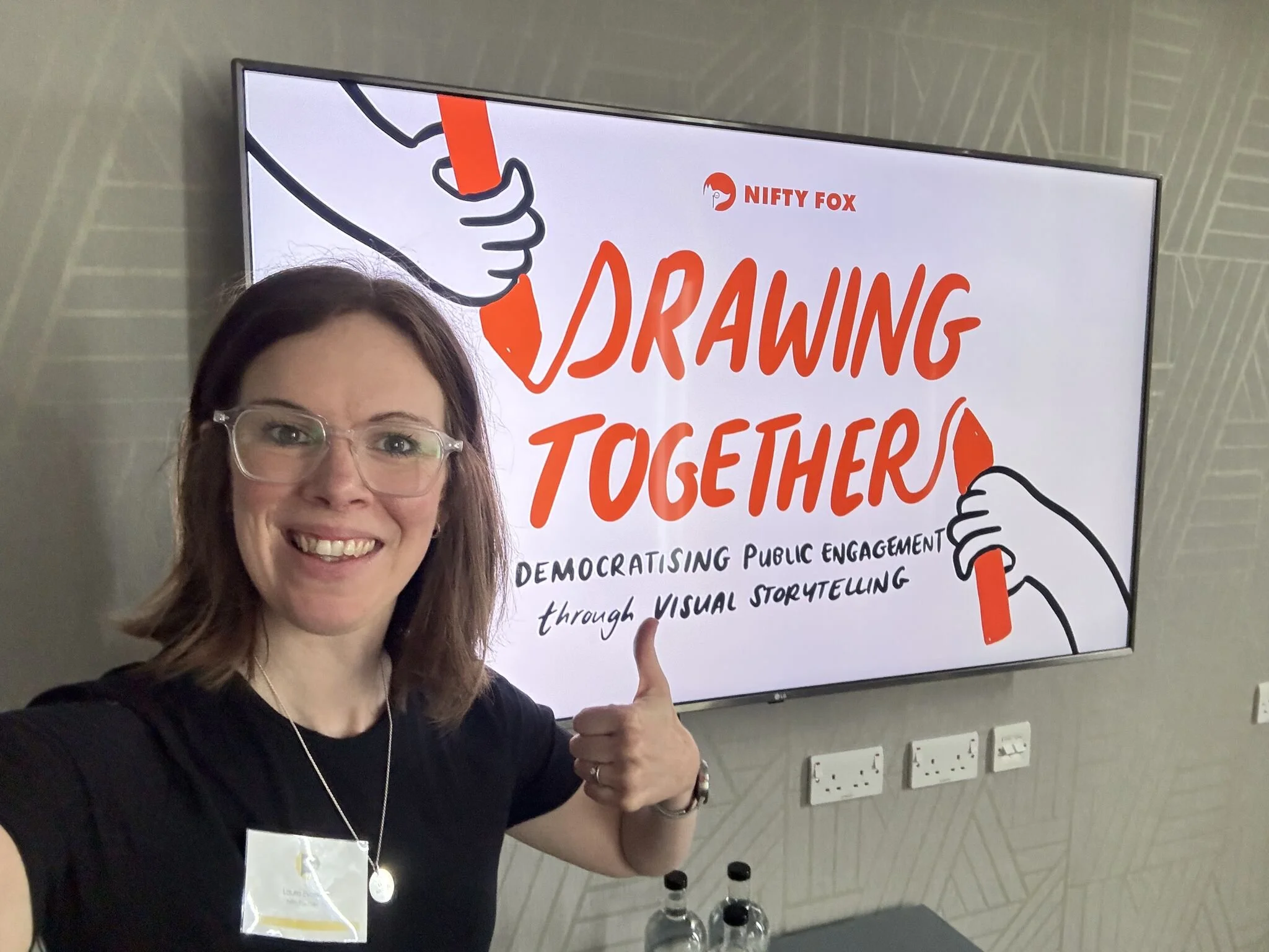 Laura standing in front of a TV screen that says 'Drawing Together'. She has her thumbs up.